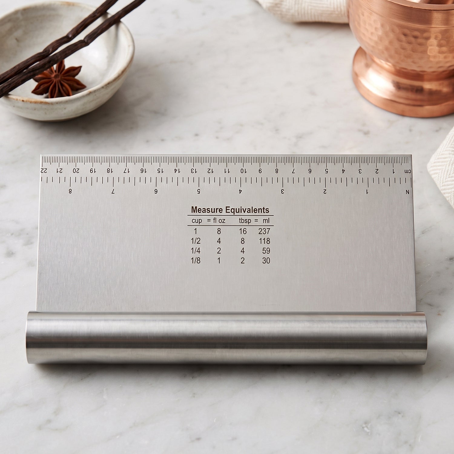 A stainless steel dough scraper with a ruler and measurement equivalents sits on a white marble surface.