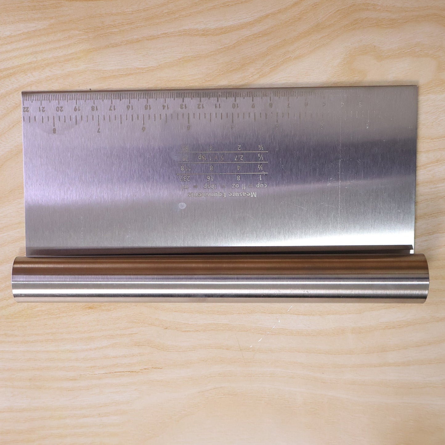 A stainless steel dough scraper with a ruler and measurement equivalents is displayed on a light wooden background.