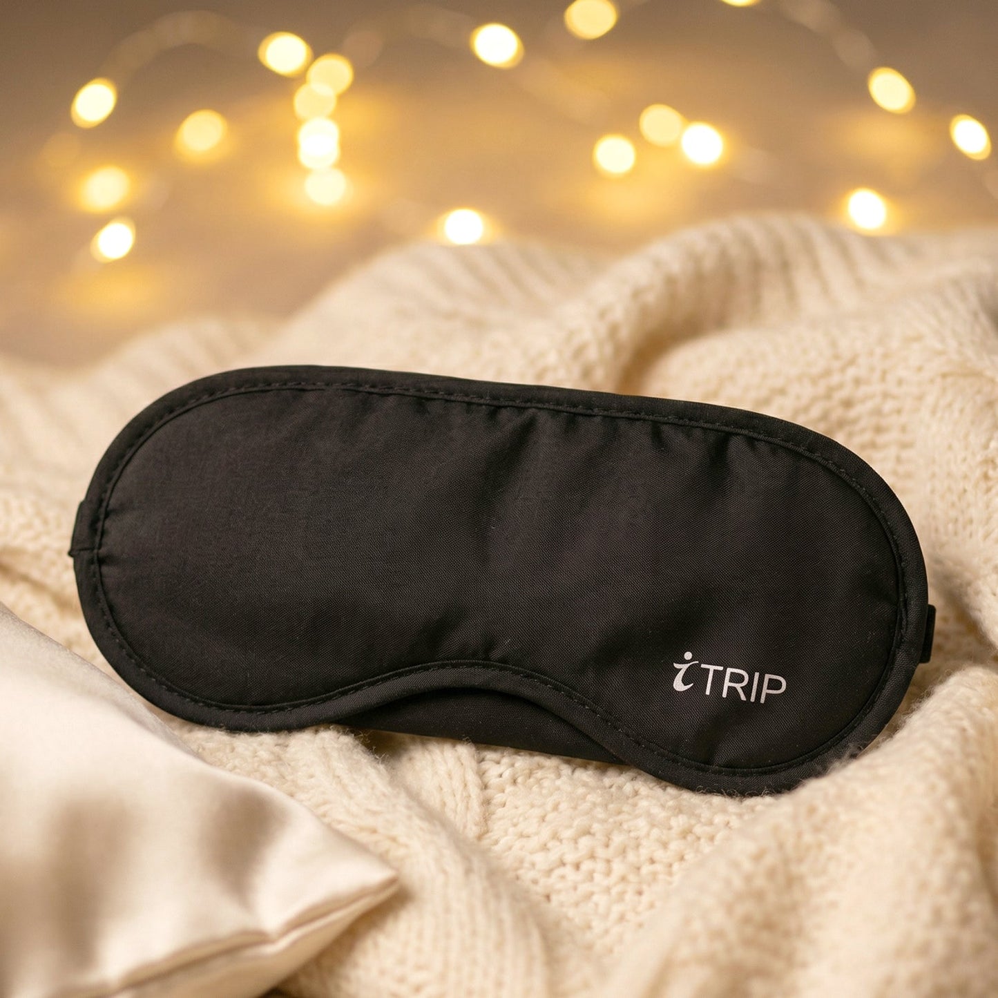 A black sleeping eye mask with the "iTRIP" logo lies on a cream cable-knit blanket, with warm fairy lights in the background.