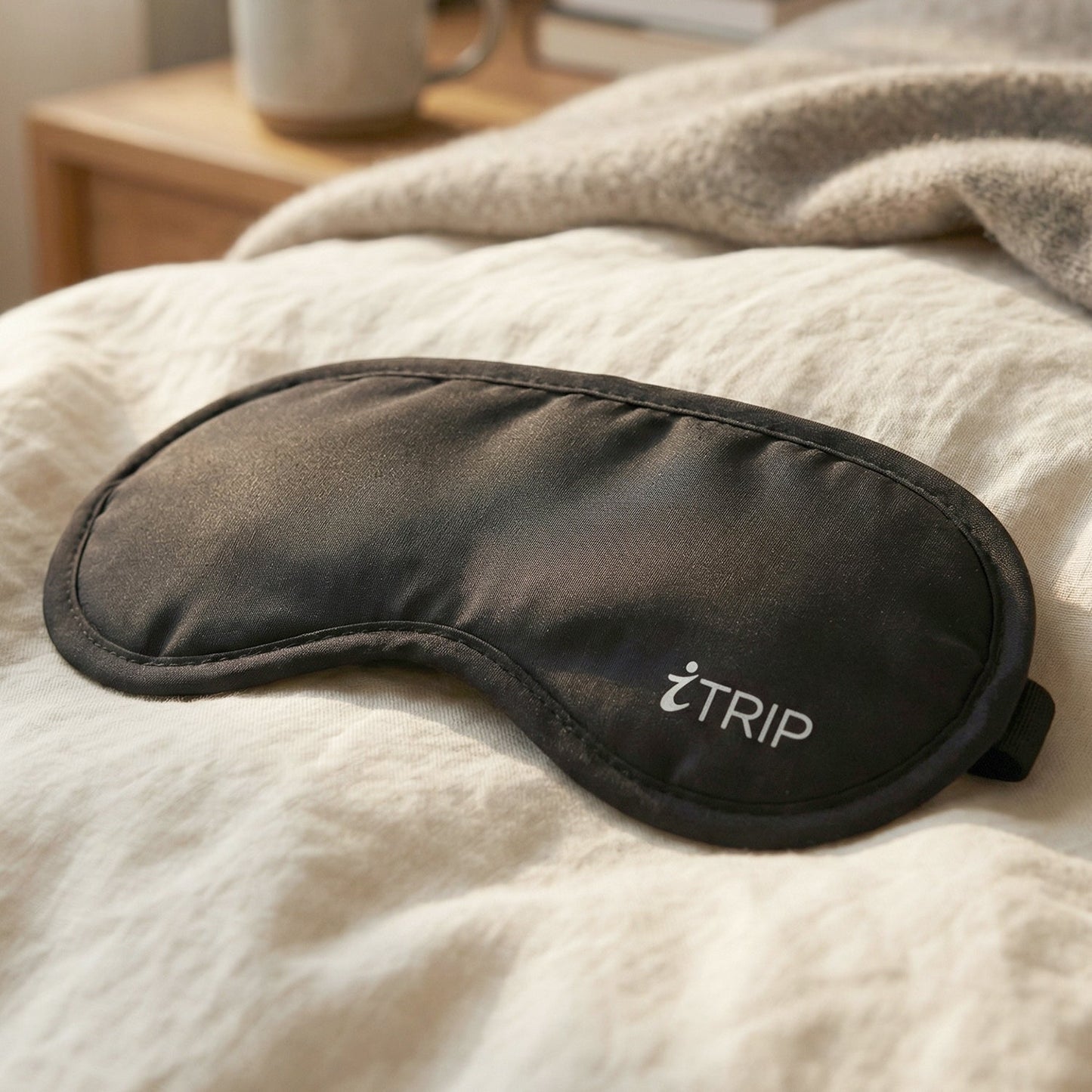 A black sleeping eye mask with the "iTRIP" logo rests on a white linen bedspread, with a blurred background of a nightstand.