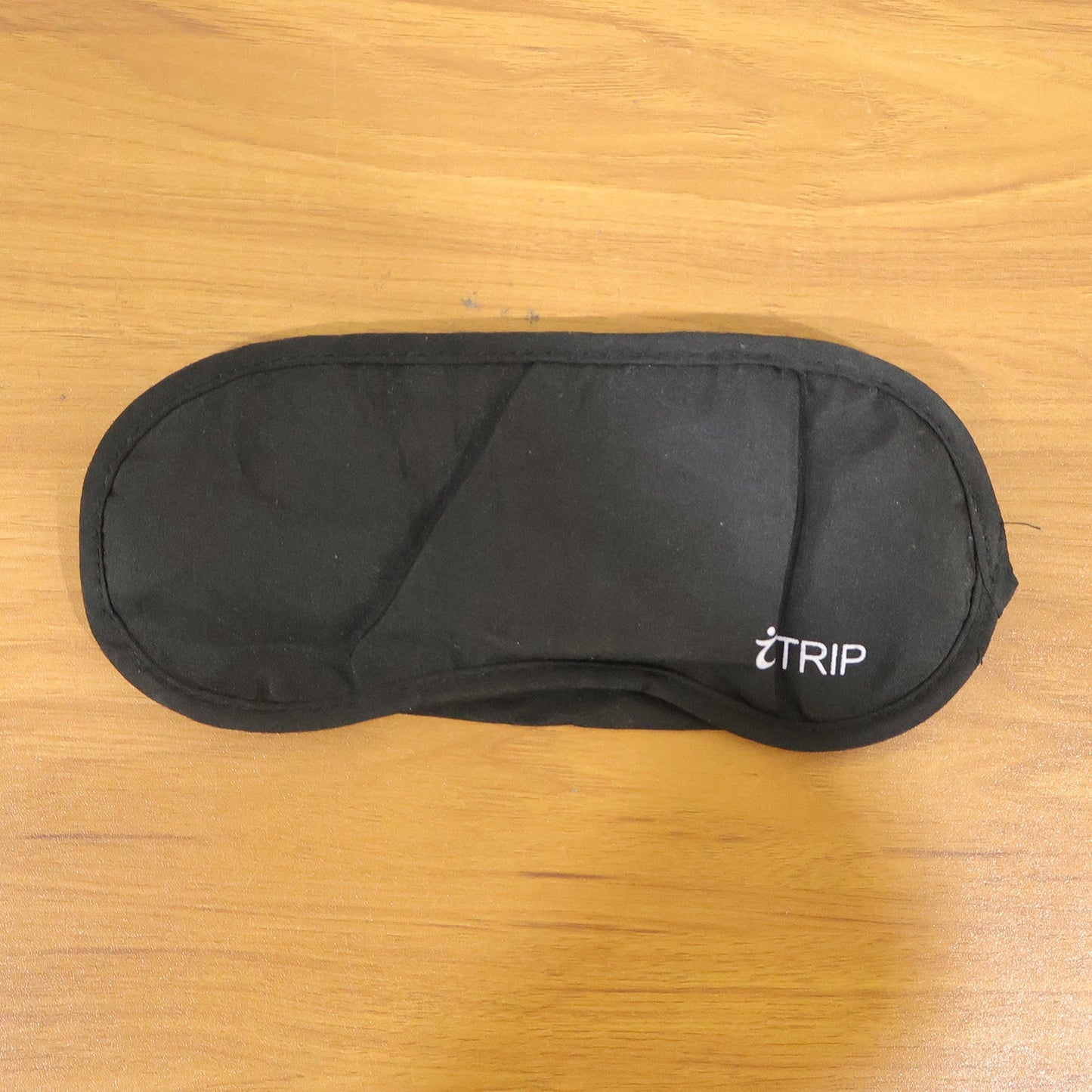 A black sleeping eye mask with the "iTRIP" logo is laid flat on a light brown wooden surface.