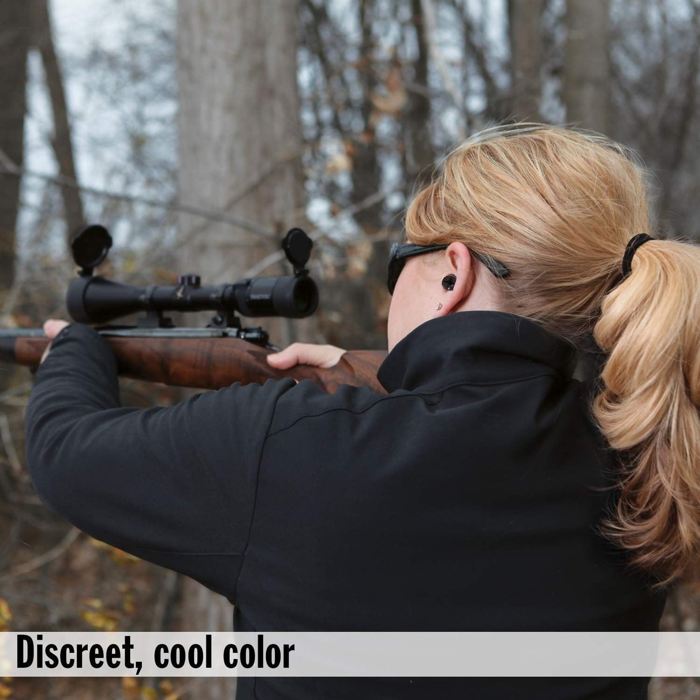 A woman with blonde hair in a ponytail aims a rifle, wearing a black earplug in her right ear.