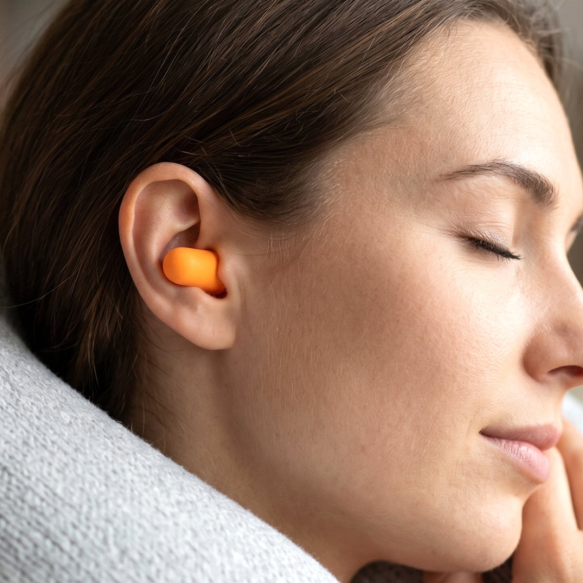 A woman with closed eyes wearing an orange earplug in her right ear, resting her head on a gray blanket.