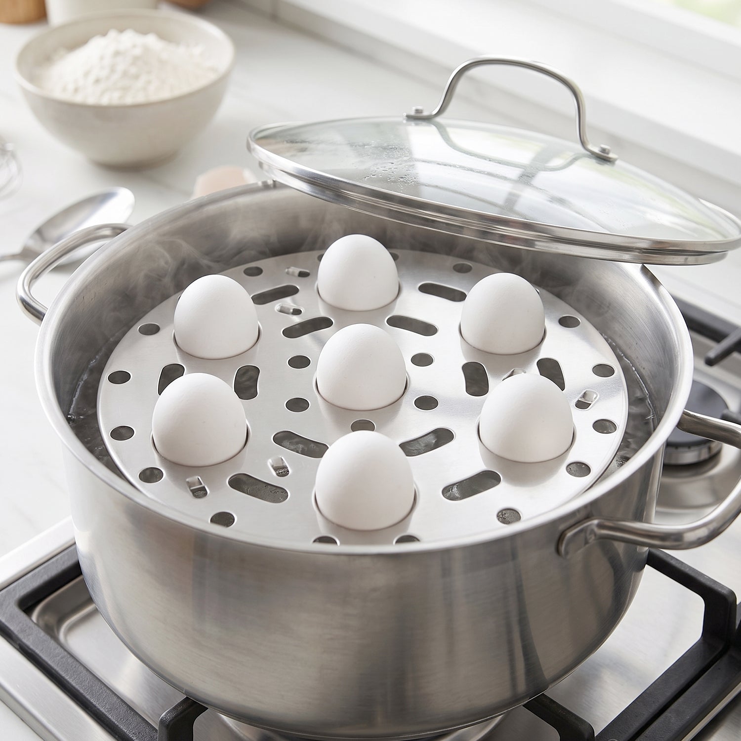 A stainless steel egg steamer rack with seven white eggs sits inside a steaming pot on a gas stove, with the pot lid propped