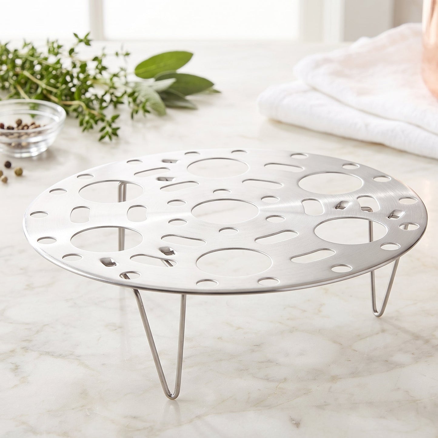 A stainless steel egg steamer rack with folding legs rests on a white marble countertop, with green herbs and a bowl of