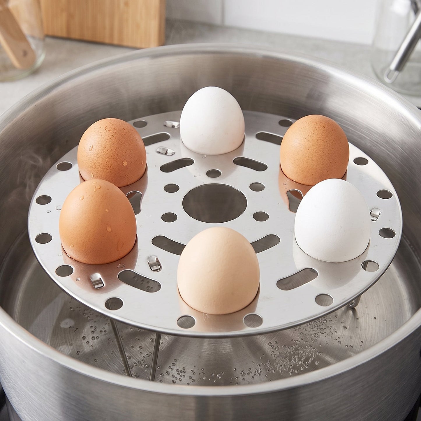 A stainless steel egg steamer rack holds six eggs, three white and three brown, inside a pot of boiling water on a stovetop.