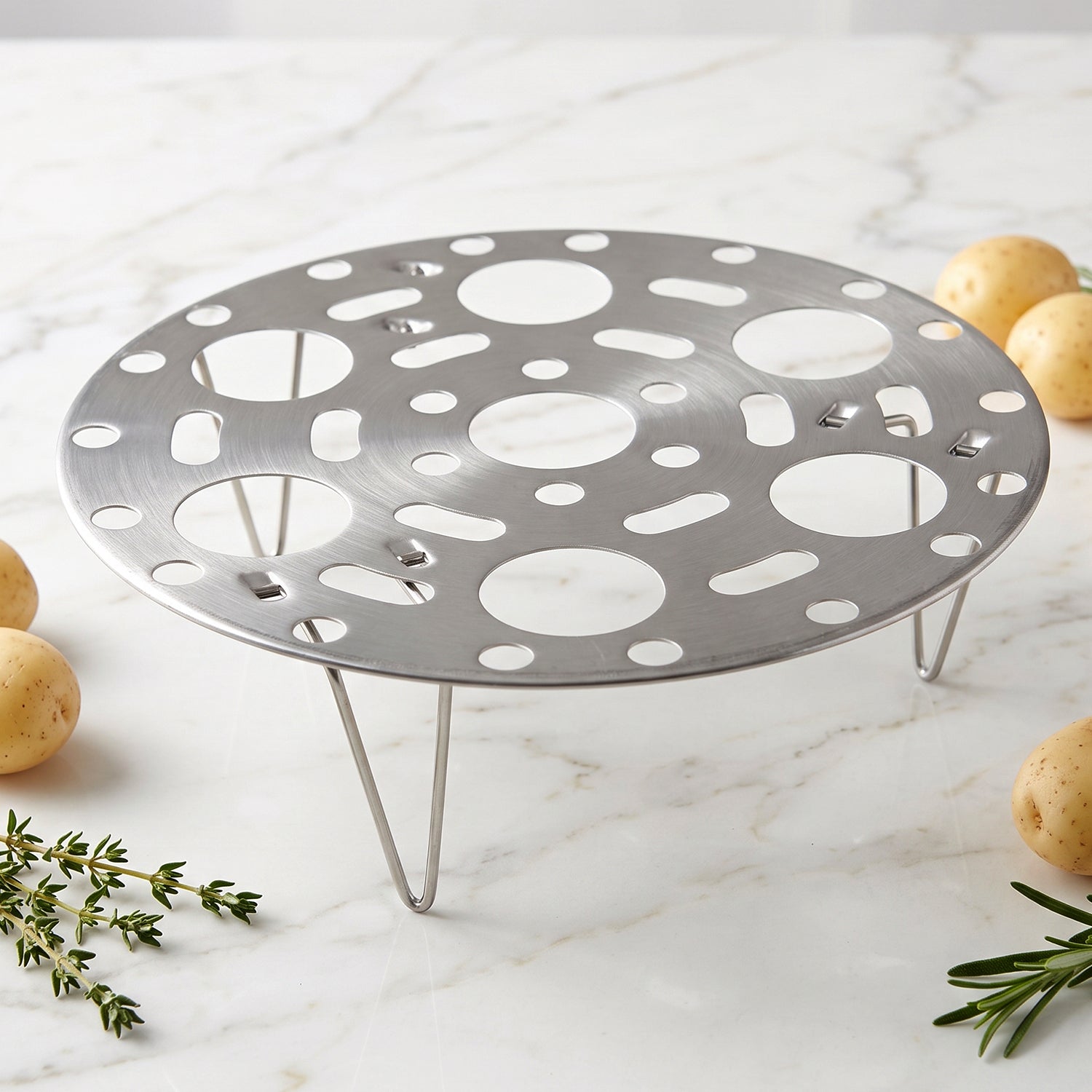A stainless steel egg steamer rack with folding legs sits on a white marble counter, surrounded by scattered small potatoes