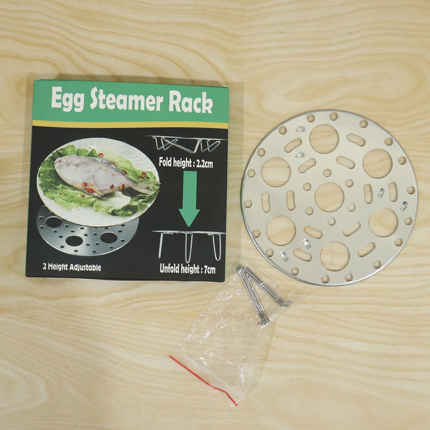 A stainless steel egg steamer rack, its packaging box, and a small plastic bag of extra legs are displayed on a light wooden