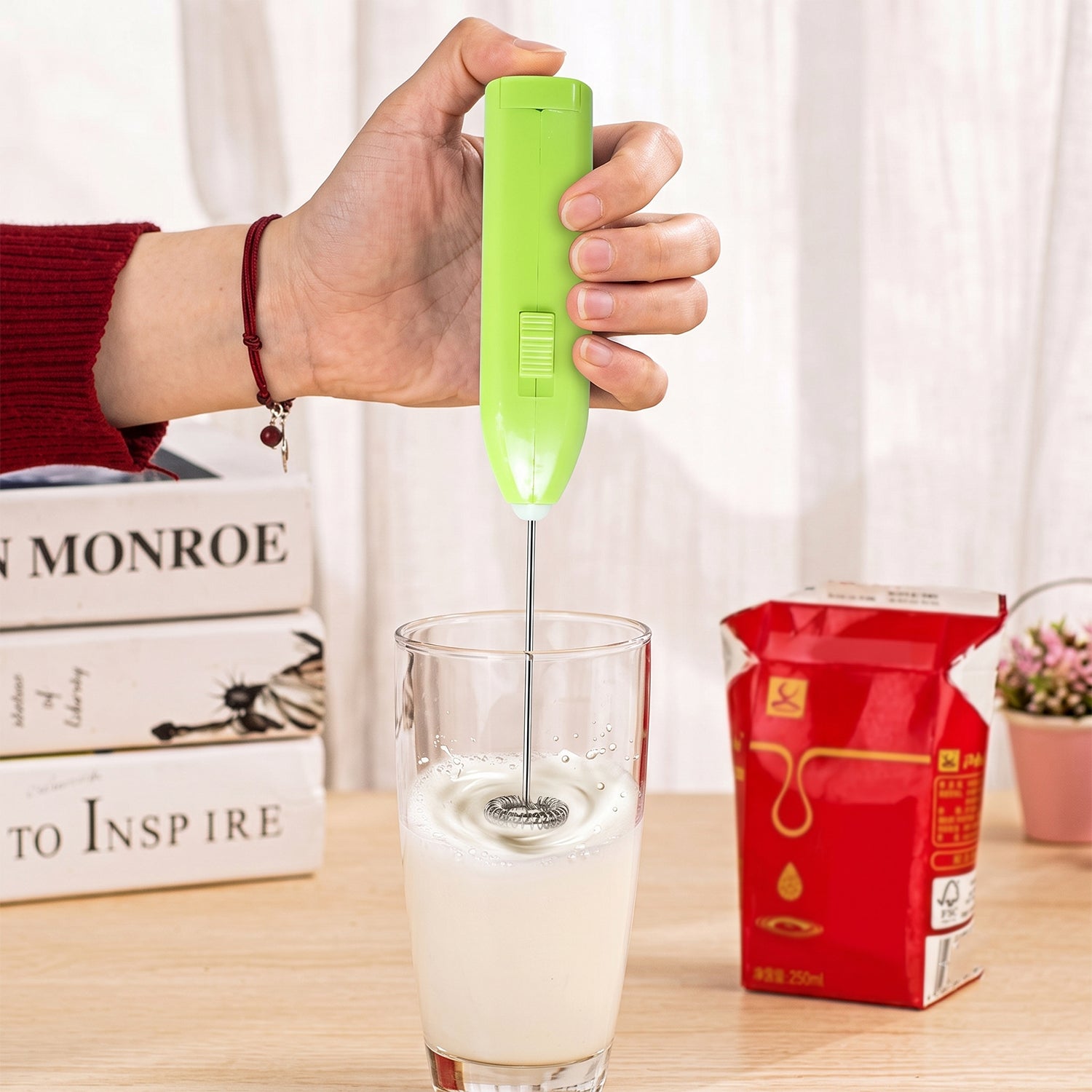 A hand holds a bright green electric frother, mixing milk in a clear glass, with books and milk carton in the background.