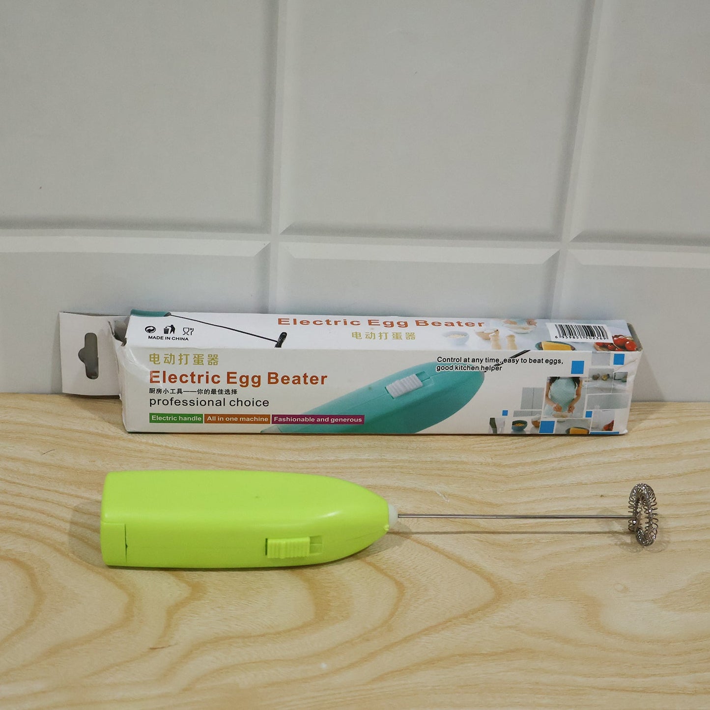 A bright green electric frother rests on a wooden floor next to its white and blue product box.