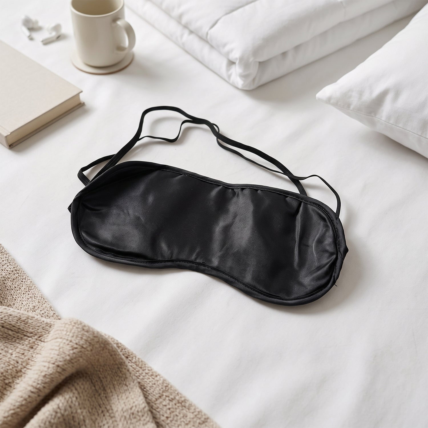 A black silk satin sleep mask lying on a white bed with a book, mug, and white bedding.