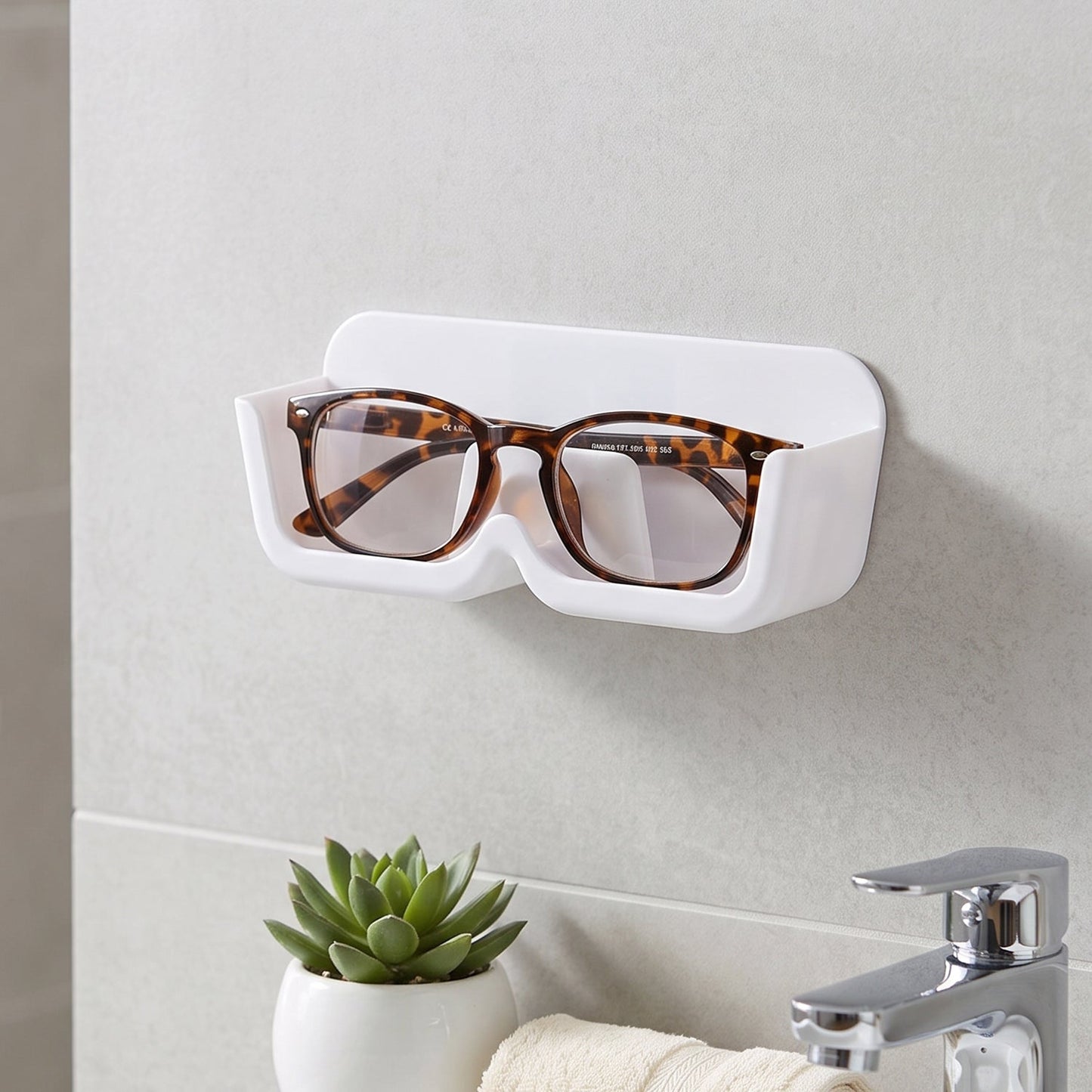 White plastic wall-mounted eyewear holder with tortoise-shell glasses, above a sink with a small green plant.