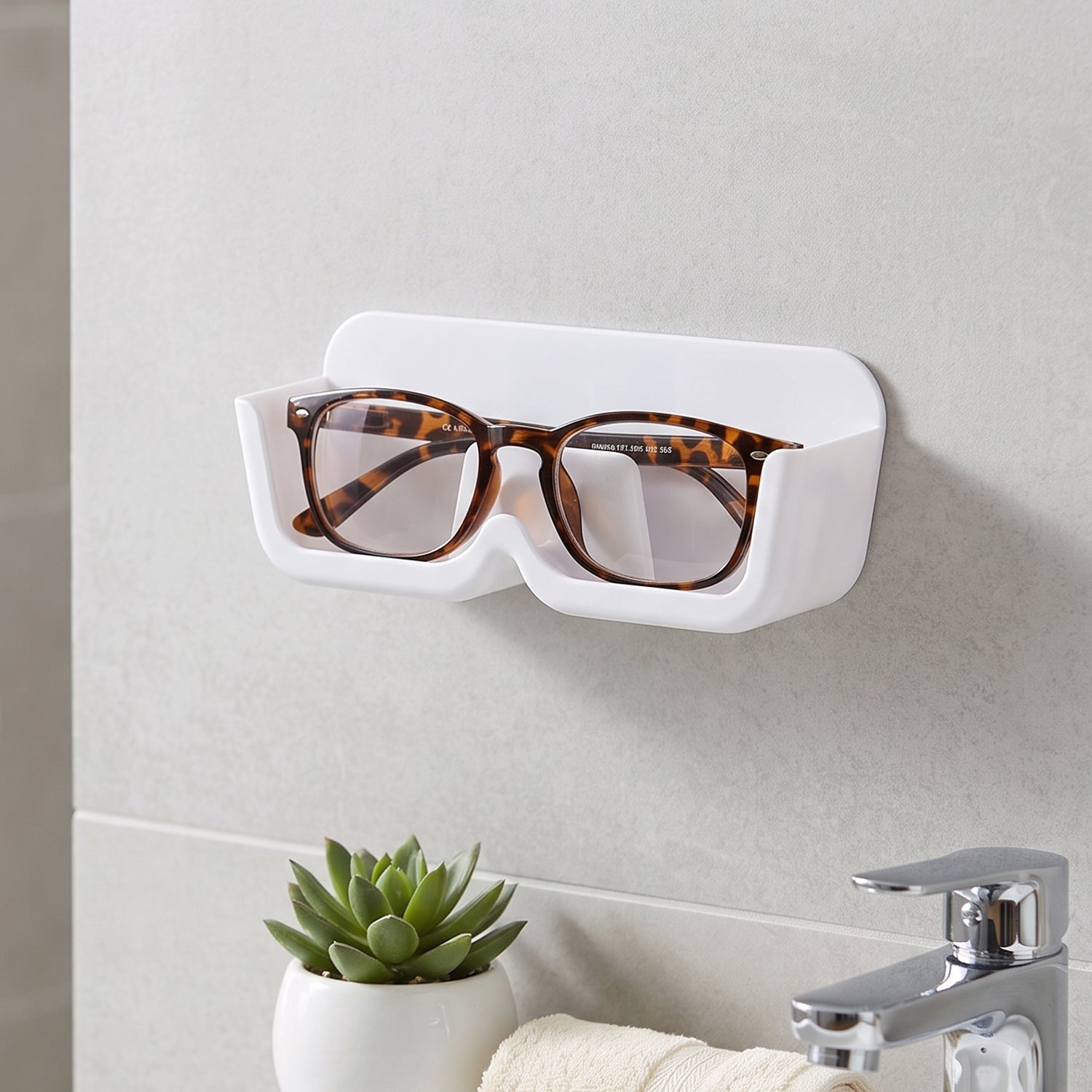 White plastic wall-mounted eyewear holder with tortoise-shell glasses, above a sink with a small green plant.