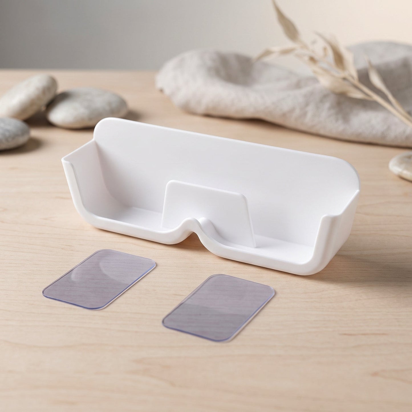 White plastic eyewear holder with two clear adhesive strips on a wooden surface, with decorative stones and fabric in the