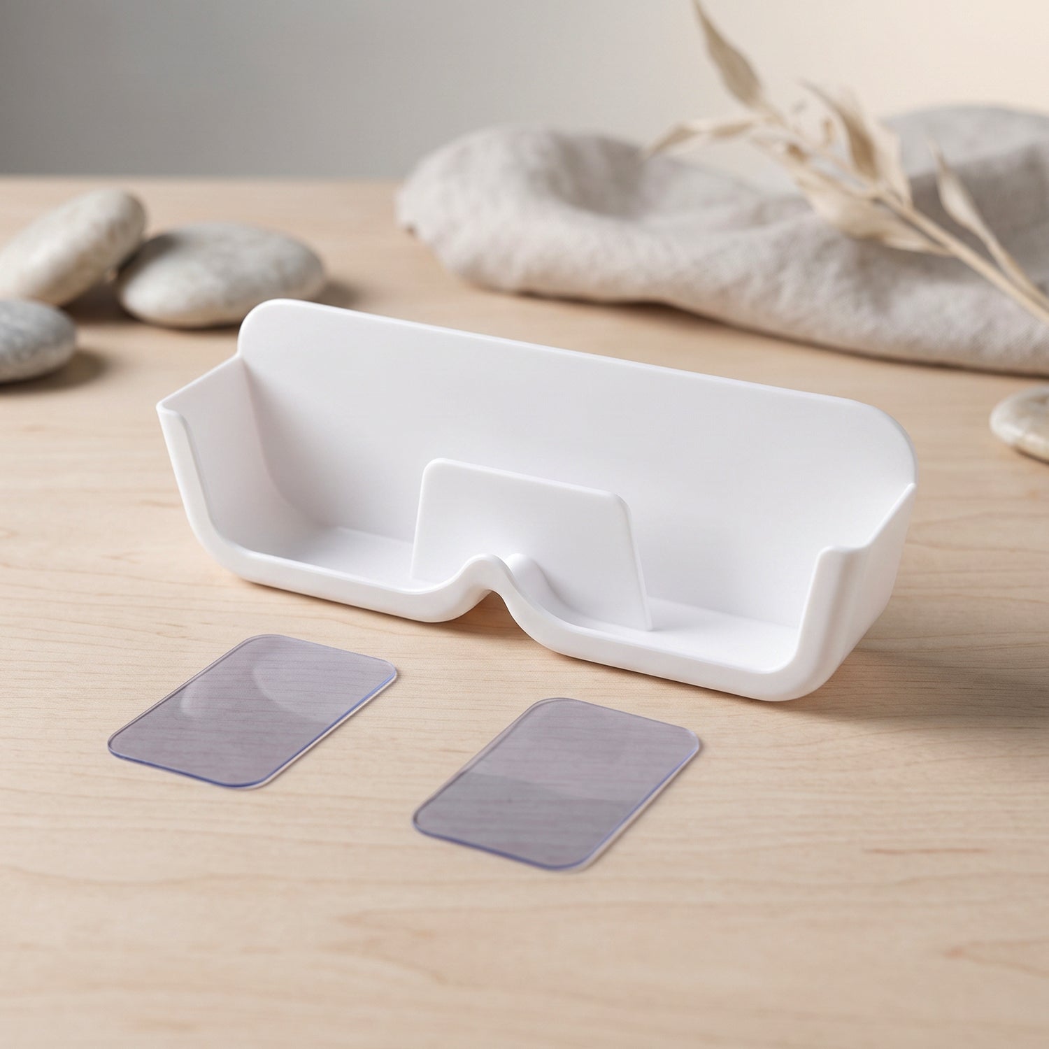 White plastic eyewear holder with two clear adhesive strips on a wooden surface, with decorative stones and fabric in the