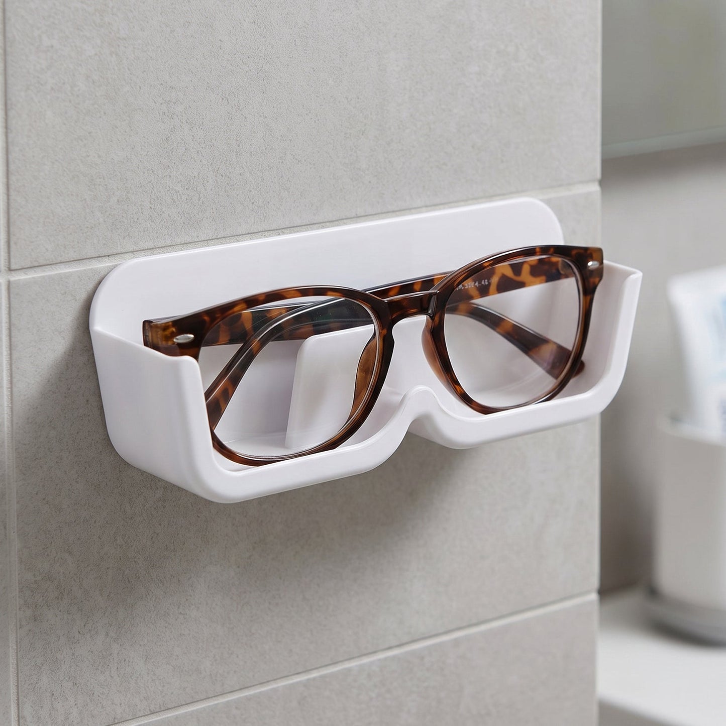 White plastic wall-mounted eyewear holder with tortoise-shell glasses, positioned on a tiled wall next to a mirror.