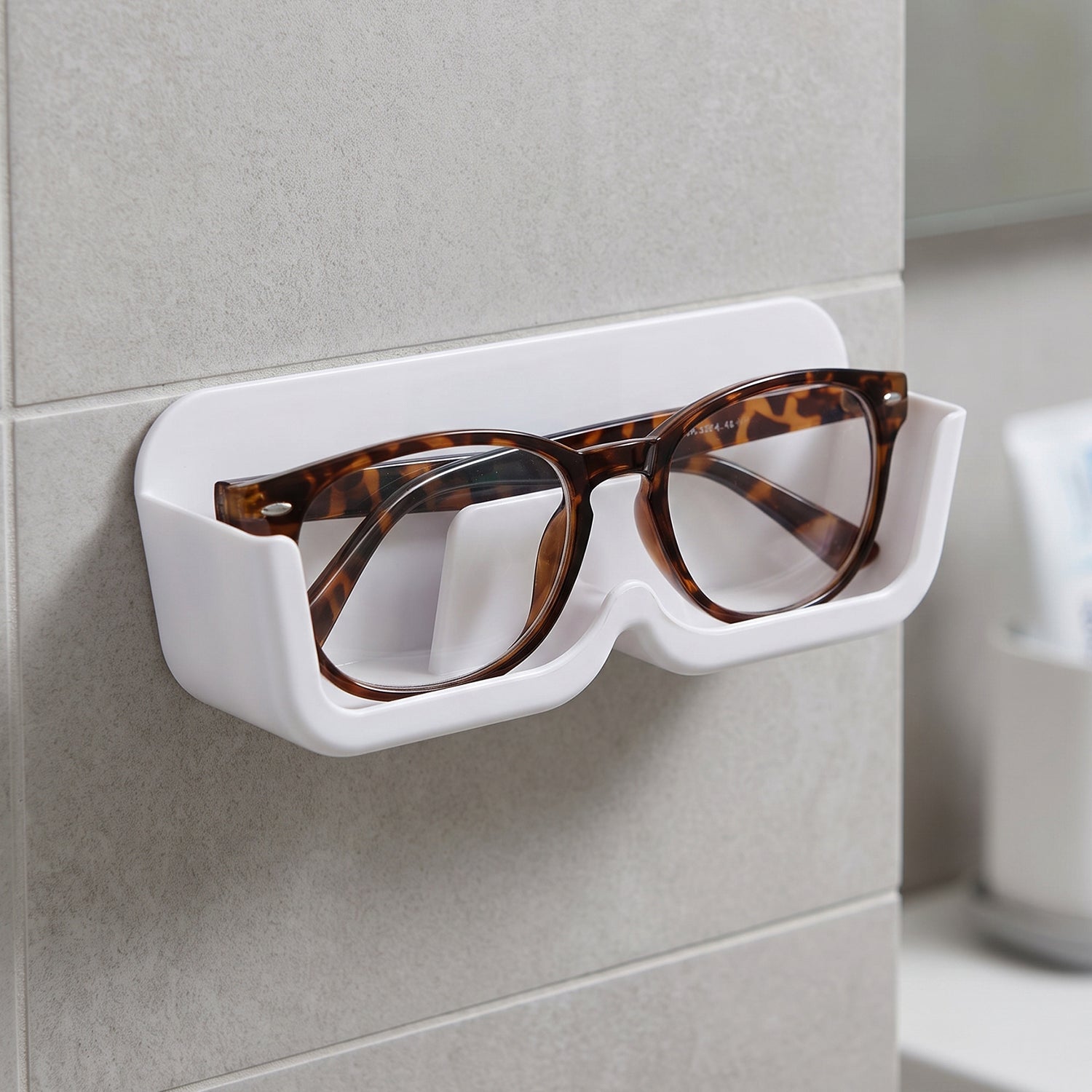 White plastic wall-mounted eyewear holder with tortoise-shell glasses, positioned on a tiled wall next to a mirror.
