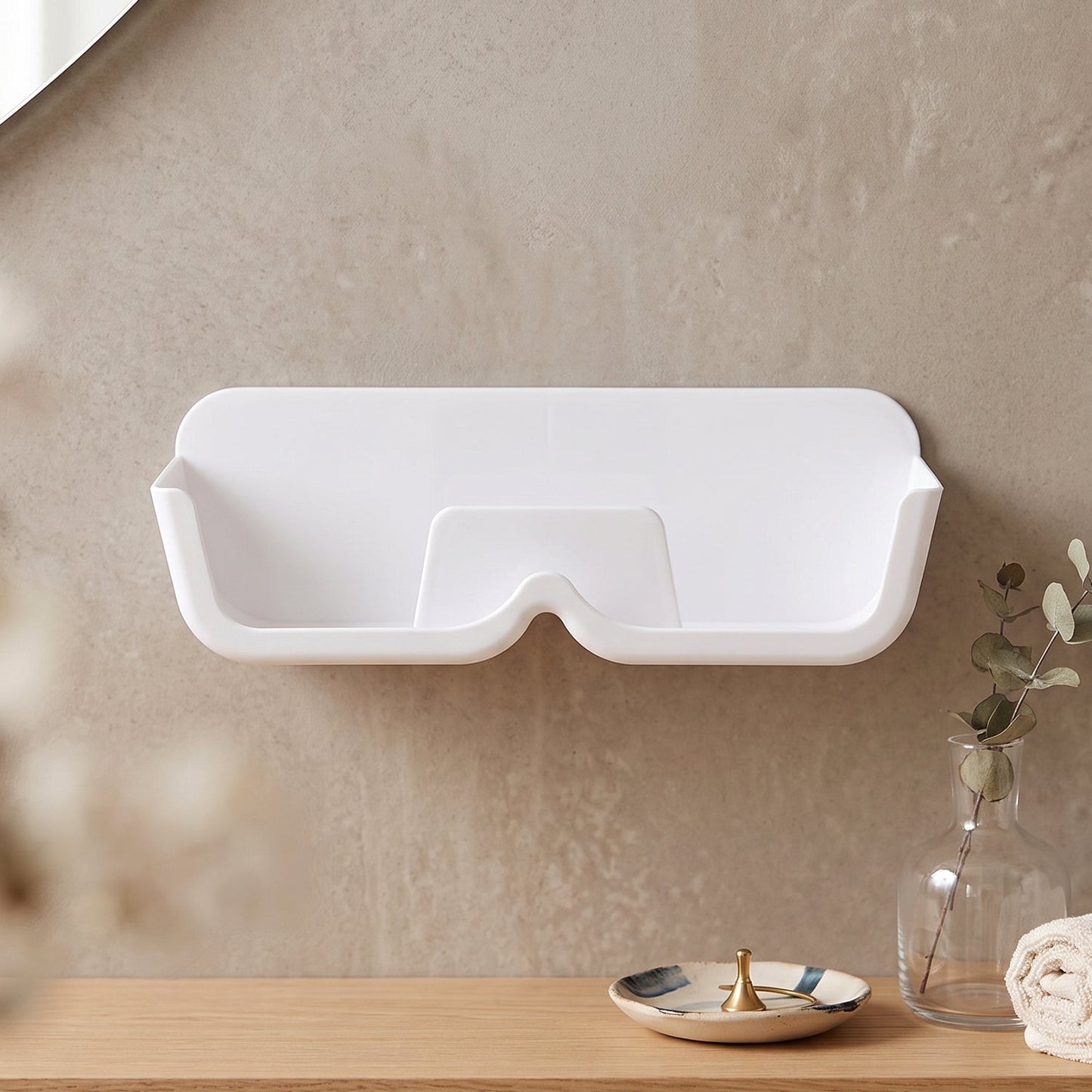 Empty white plastic wall-mounted eyewear holder on a light brown wall, with a vase and towel on a wooden surface below.