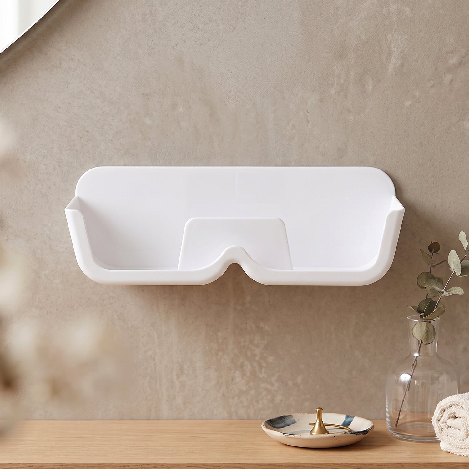 Empty white plastic wall-mounted eyewear holder on a light brown wall, with a vase and towel on a wooden surface below.