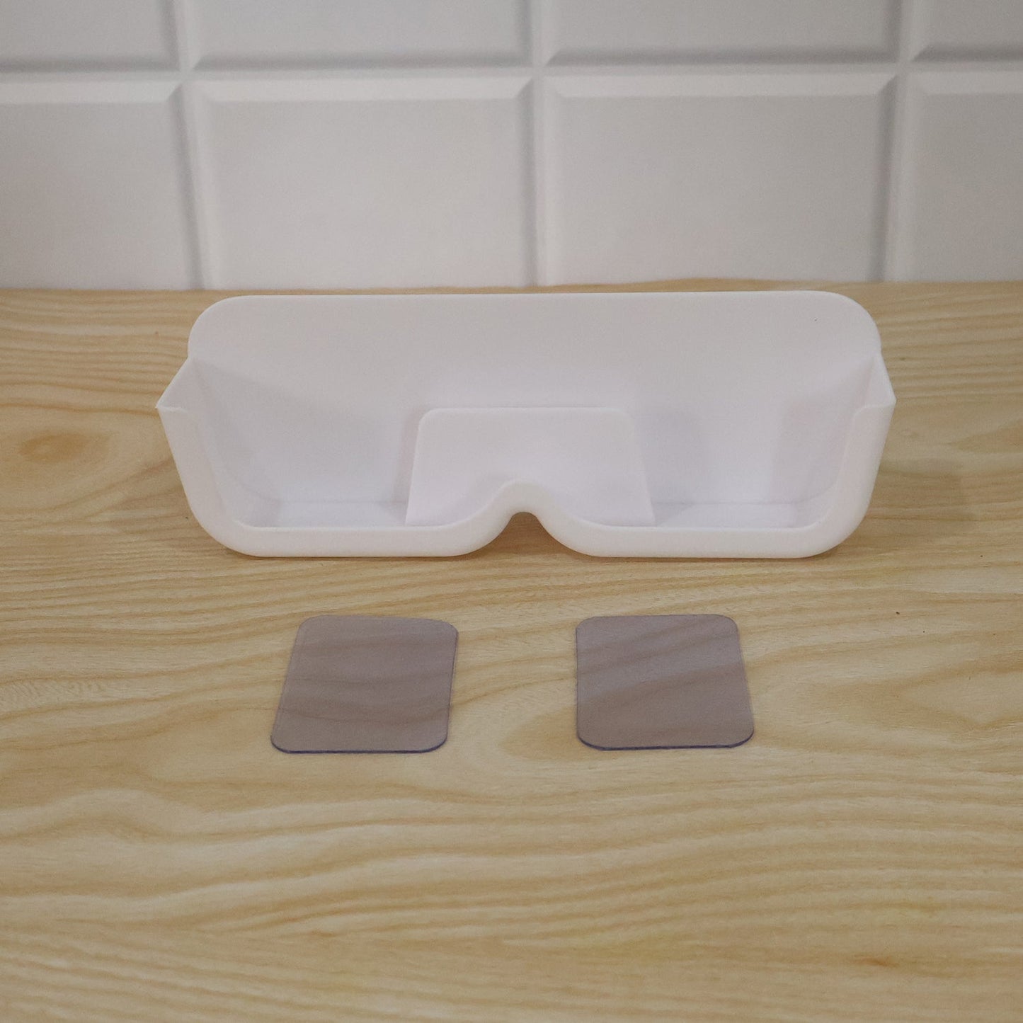 White plastic eyewear holder with two clear adhesive strips on a light wooden surface, against a white tiled wall.