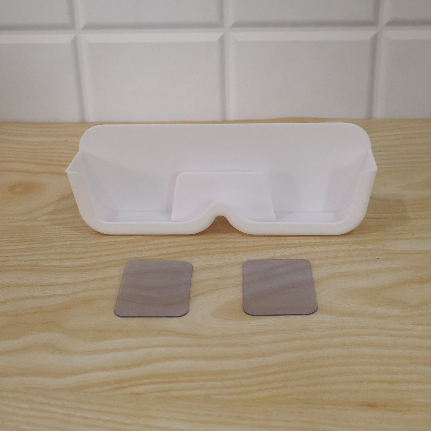 White plastic eyewear holder with two clear adhesive strips on a light wooden surface, against a white tiled wall.