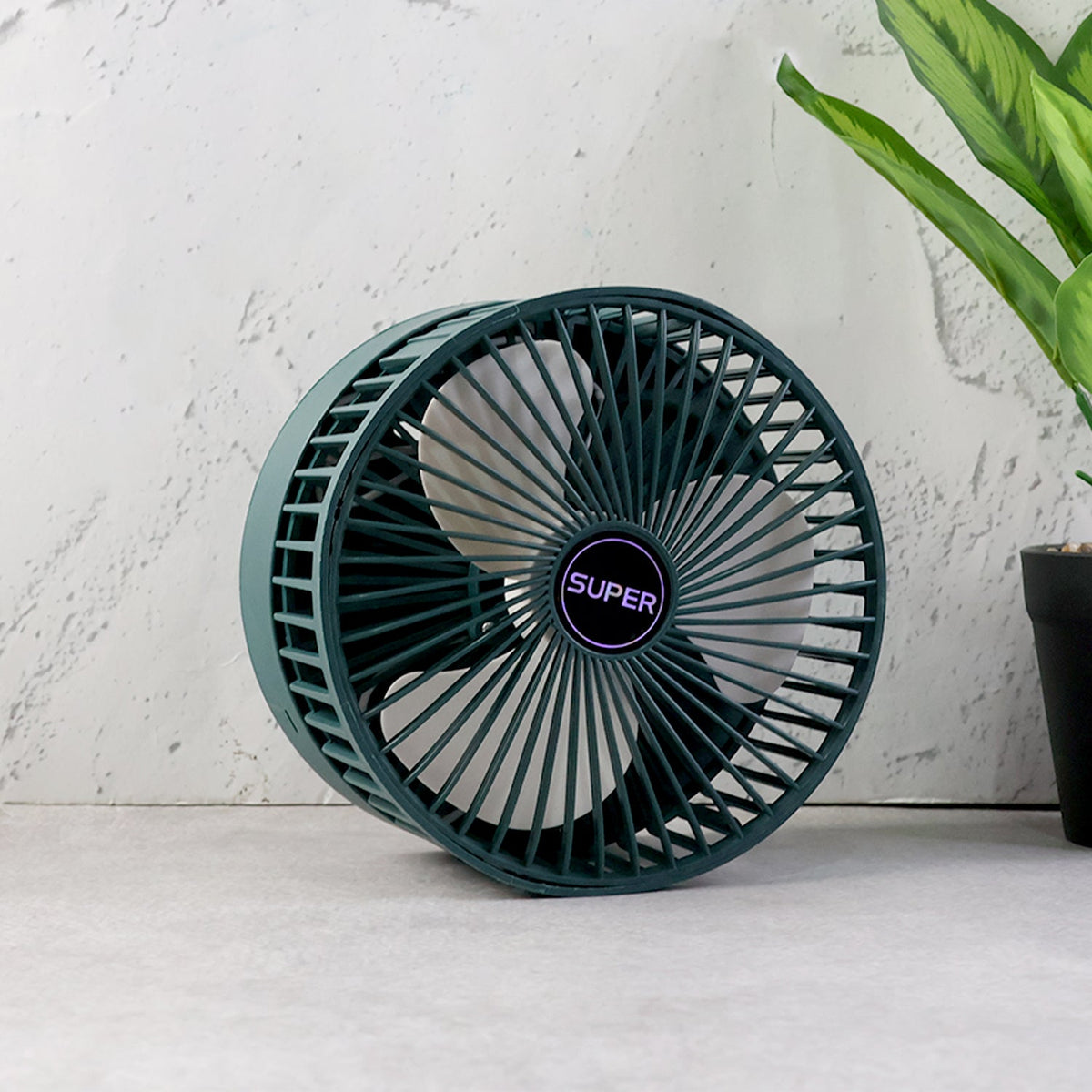 A dark green telescopic foldable mini USB desk fan in its compact, folded state, on a grey surface next to a plant.