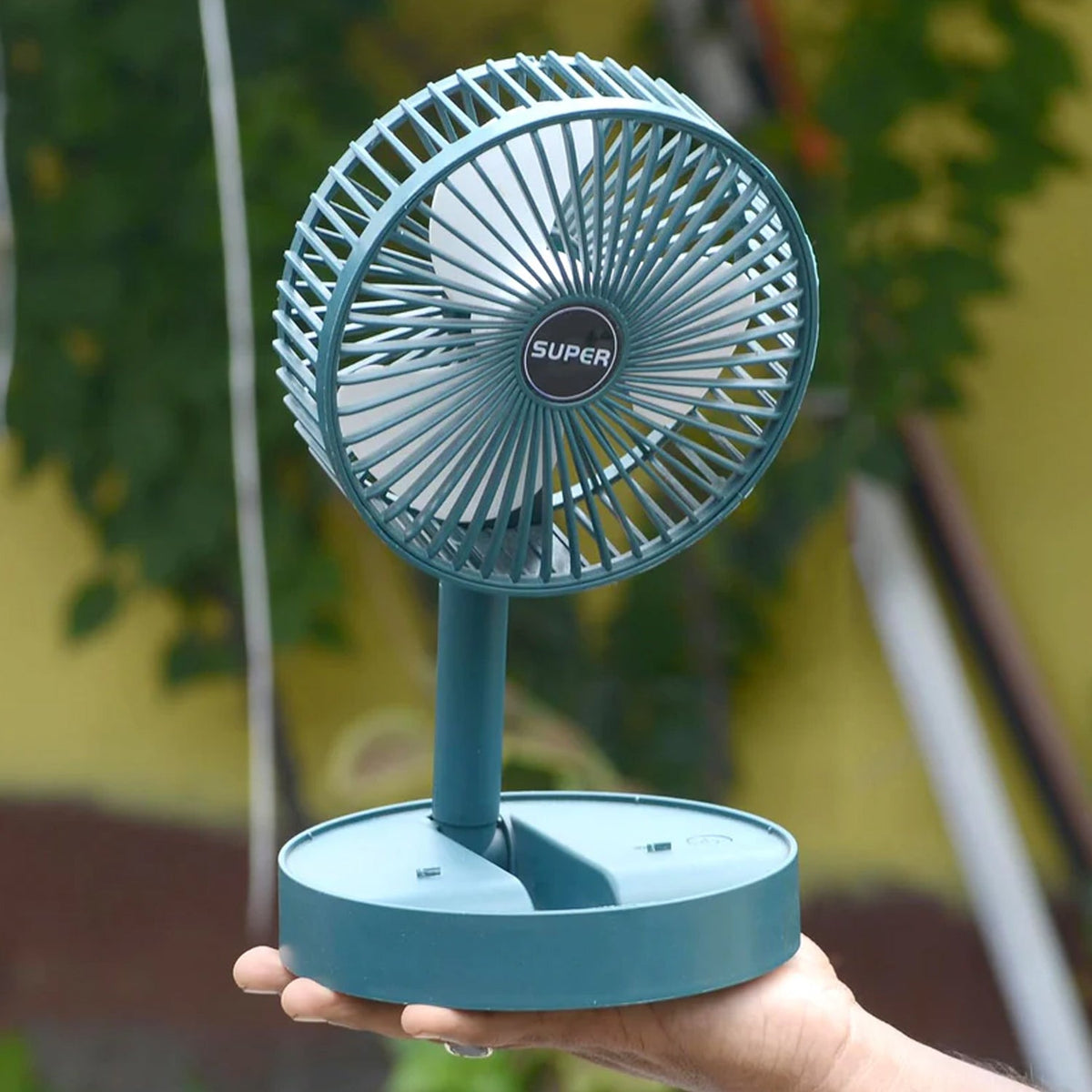 A hand holds a dark green telescopic foldable mini USB desk fan, extended, against an outdoor background.