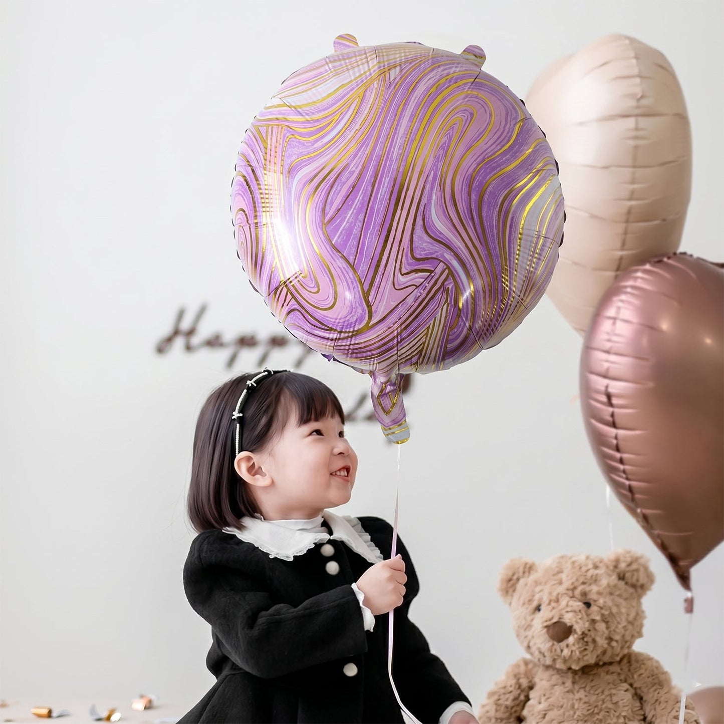 A young girl in a black dress holds a purple and gold marble foil balloon next to a teddy bear.