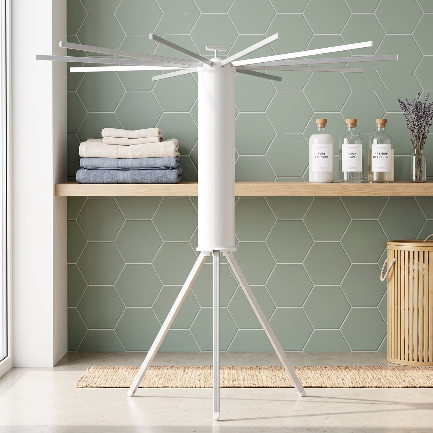 White tripod clothes drying rack in a laundry room with green hexagonal tiles, folded towels, and bottles on a shelf.