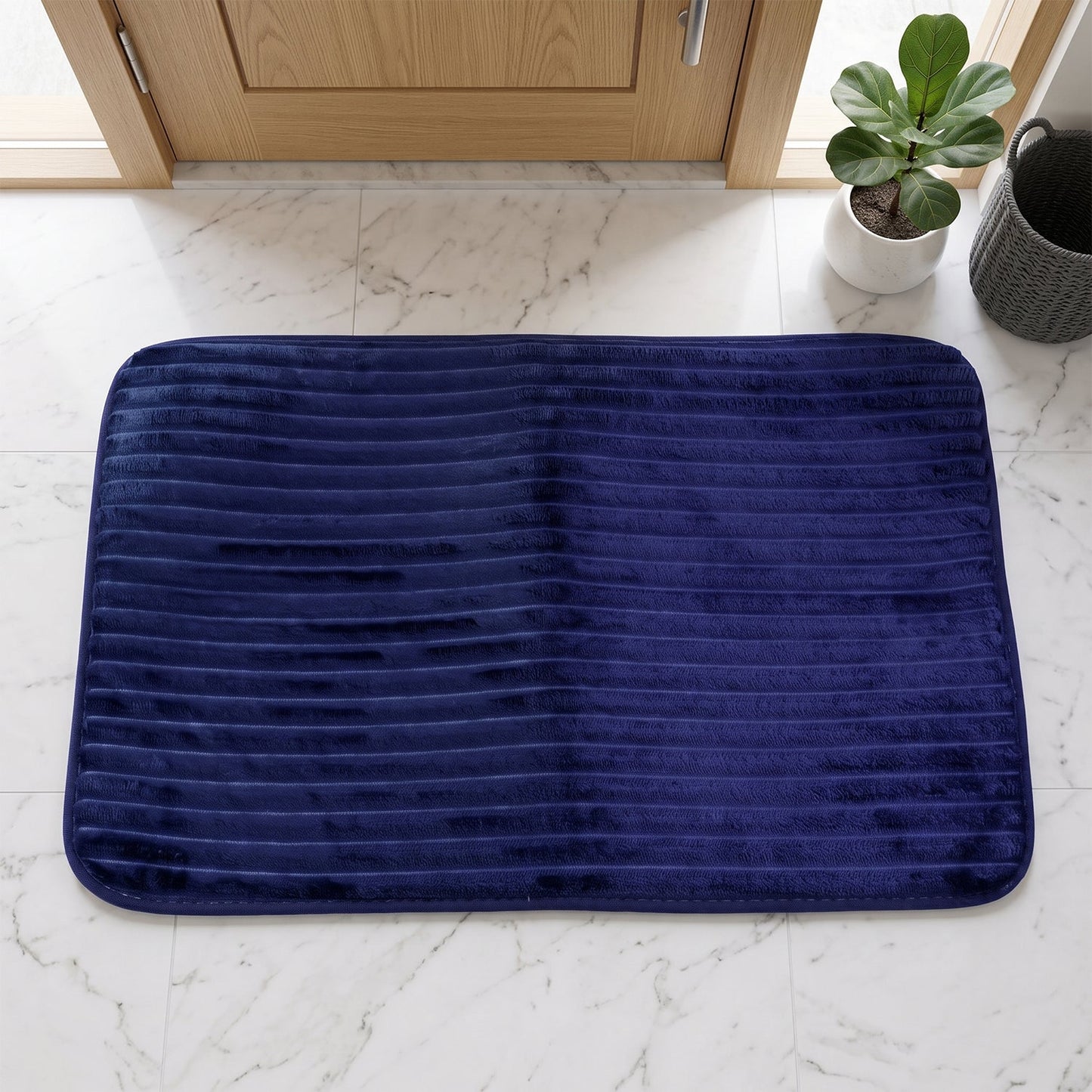 A navy blue ribbed memory foam bath mat lies on a white marble floor in front of a light wood door.