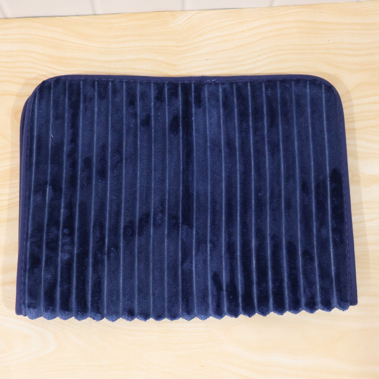A navy blue ribbed memory foam bath mat is folded in half, resting on a light wooden surface.