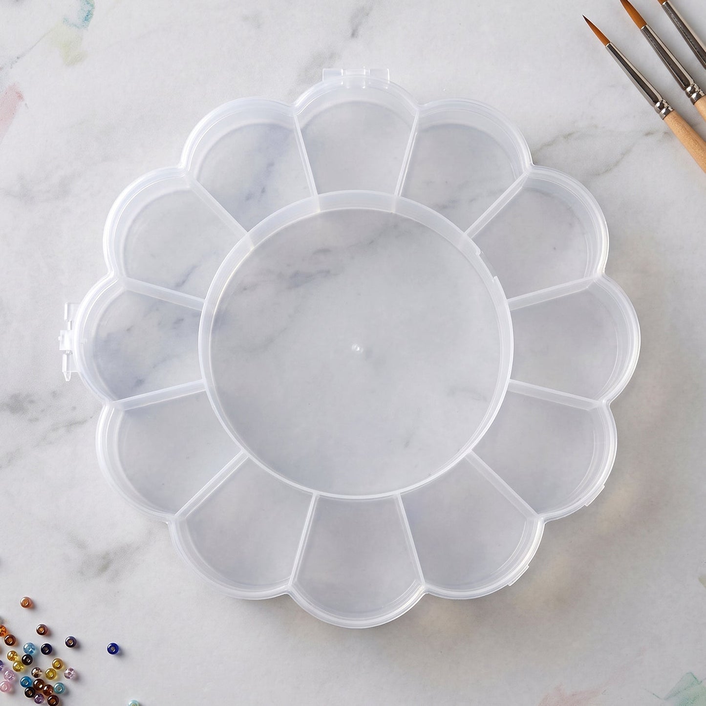 Empty clear flower-shaped plastic organizer box with 13 compartments on a white marble surface with paintbrushes nearby.