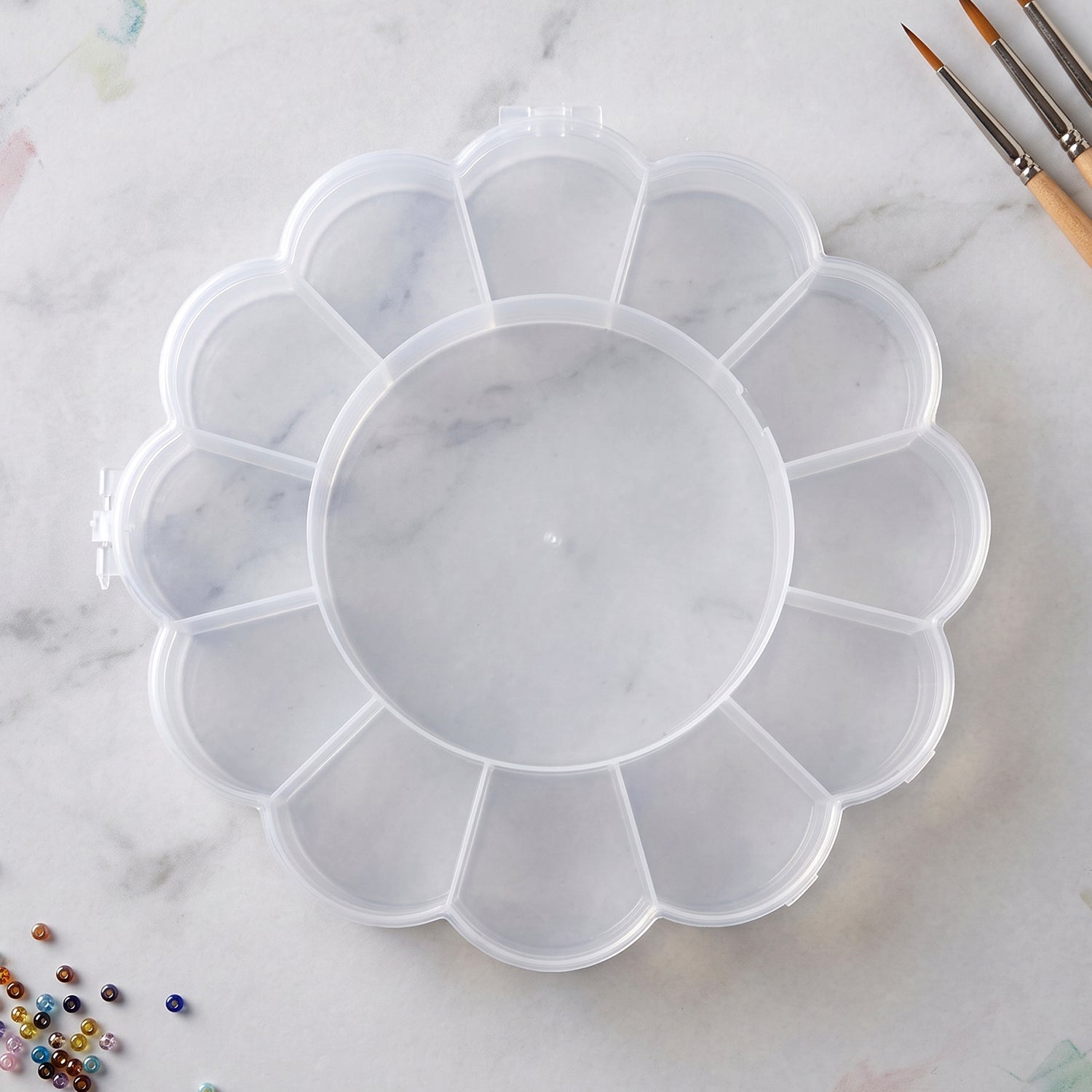 Empty clear flower-shaped plastic organizer box with 13 compartments on a white marble surface with paintbrushes nearby.