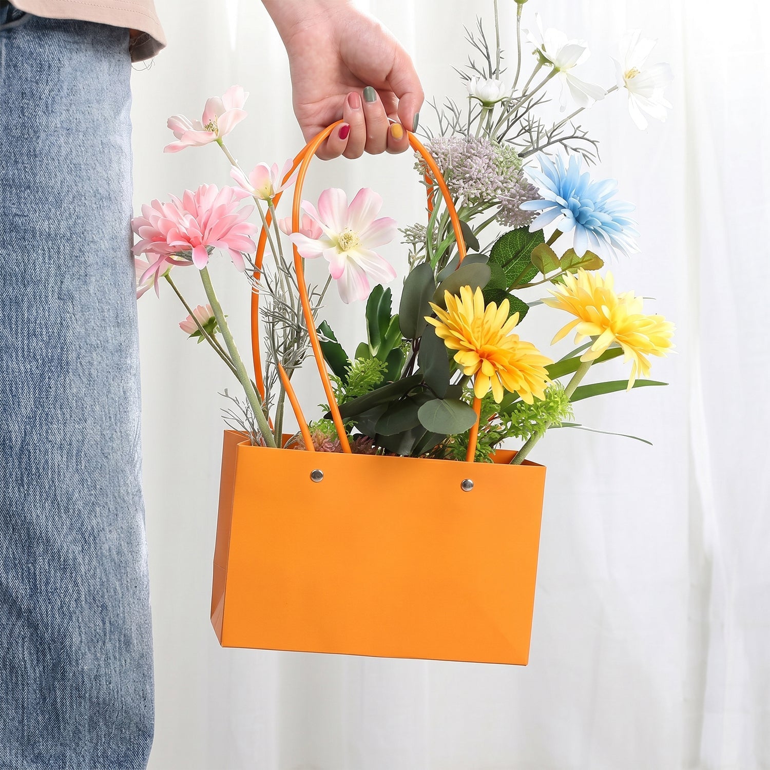 A hand holds an orange rectangular gift bag with round handles, filled with a vibrant mix of colorful artificial flowers,
