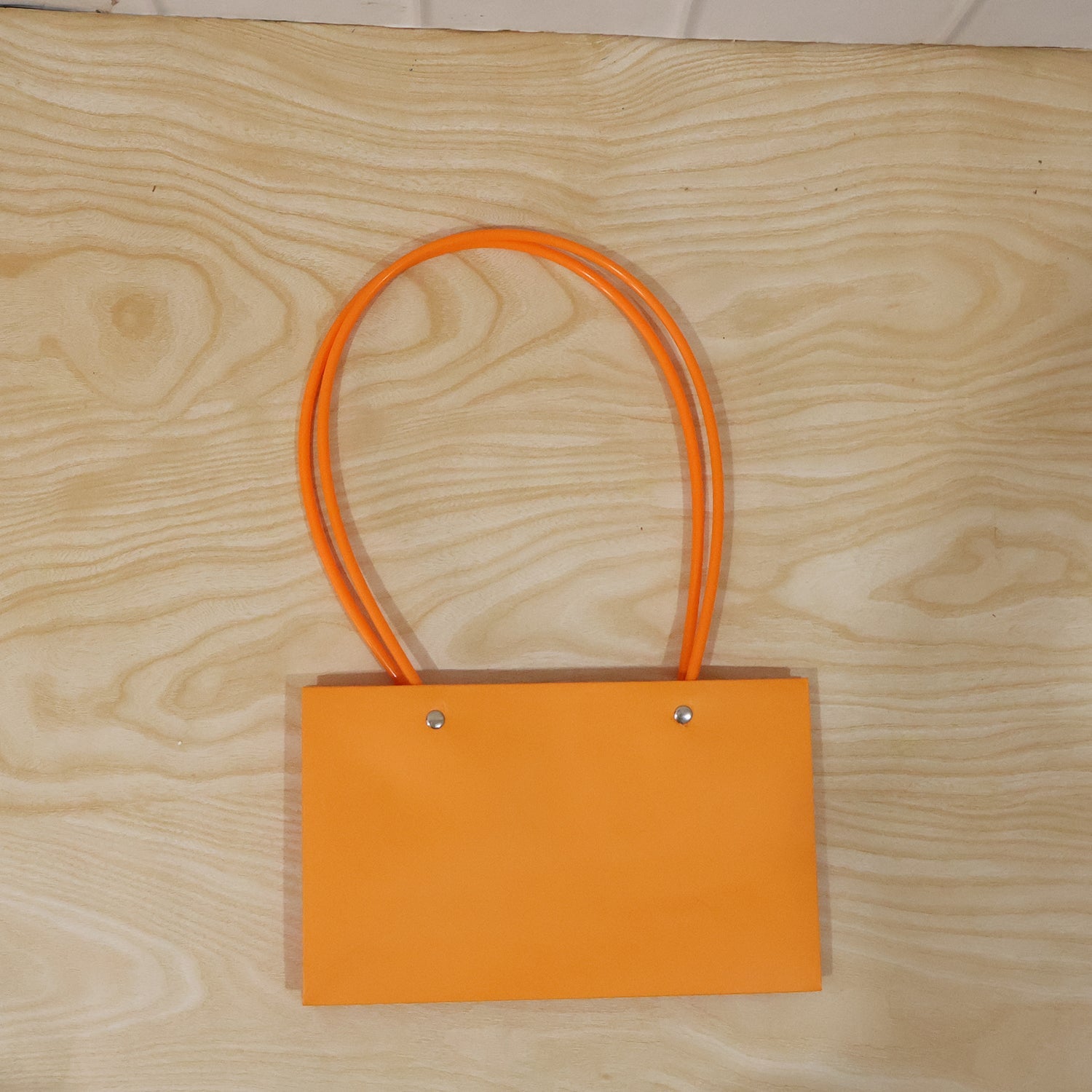 A glossy orange rectangular gift bag with thin, round orange handles is centered on a light wooden background.