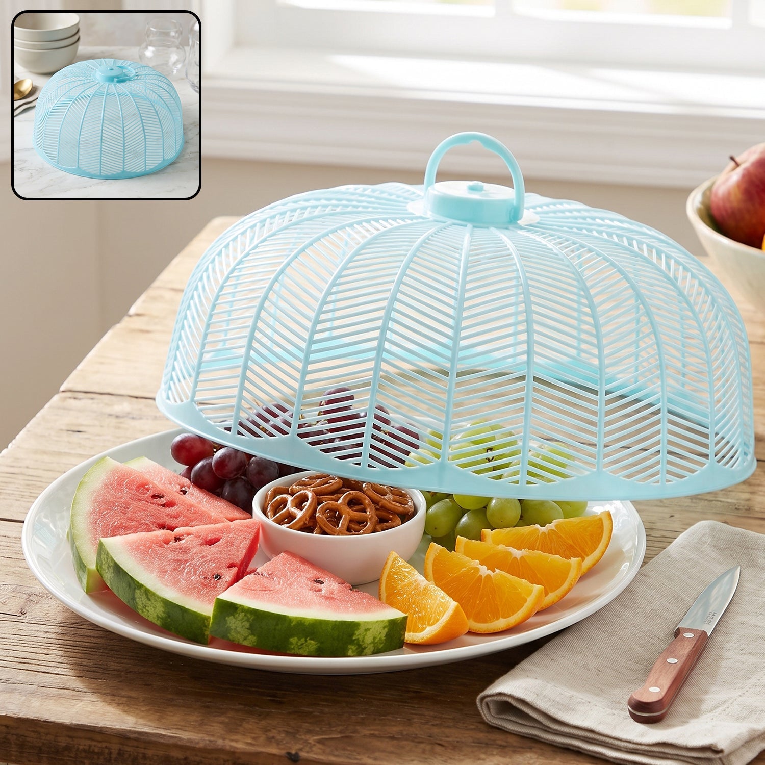 Light blue plastic mesh food cover dome over a white plate with watermelon, oranges, grapes, and pretzels.