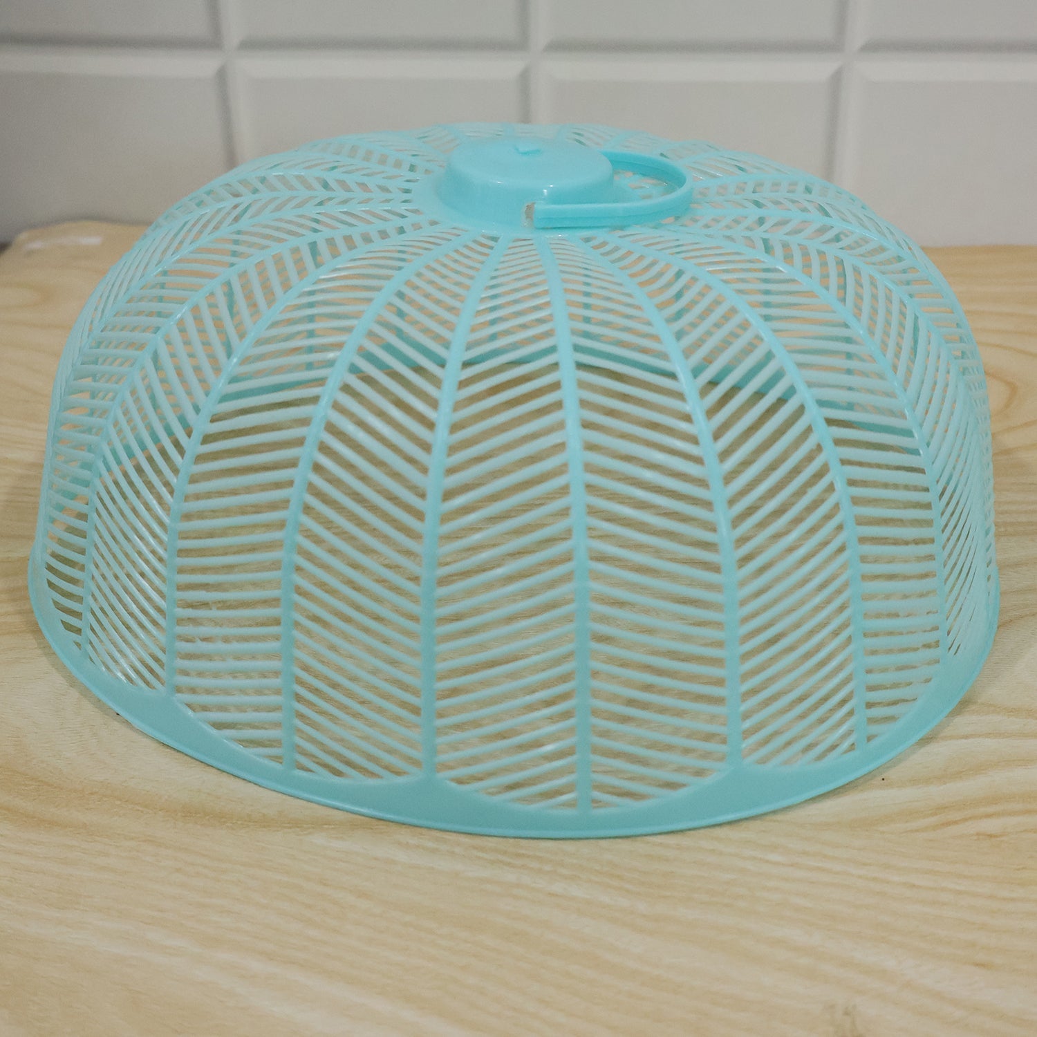 Light blue plastic mesh food cover dome on a light wooden surface with a white tiled background.