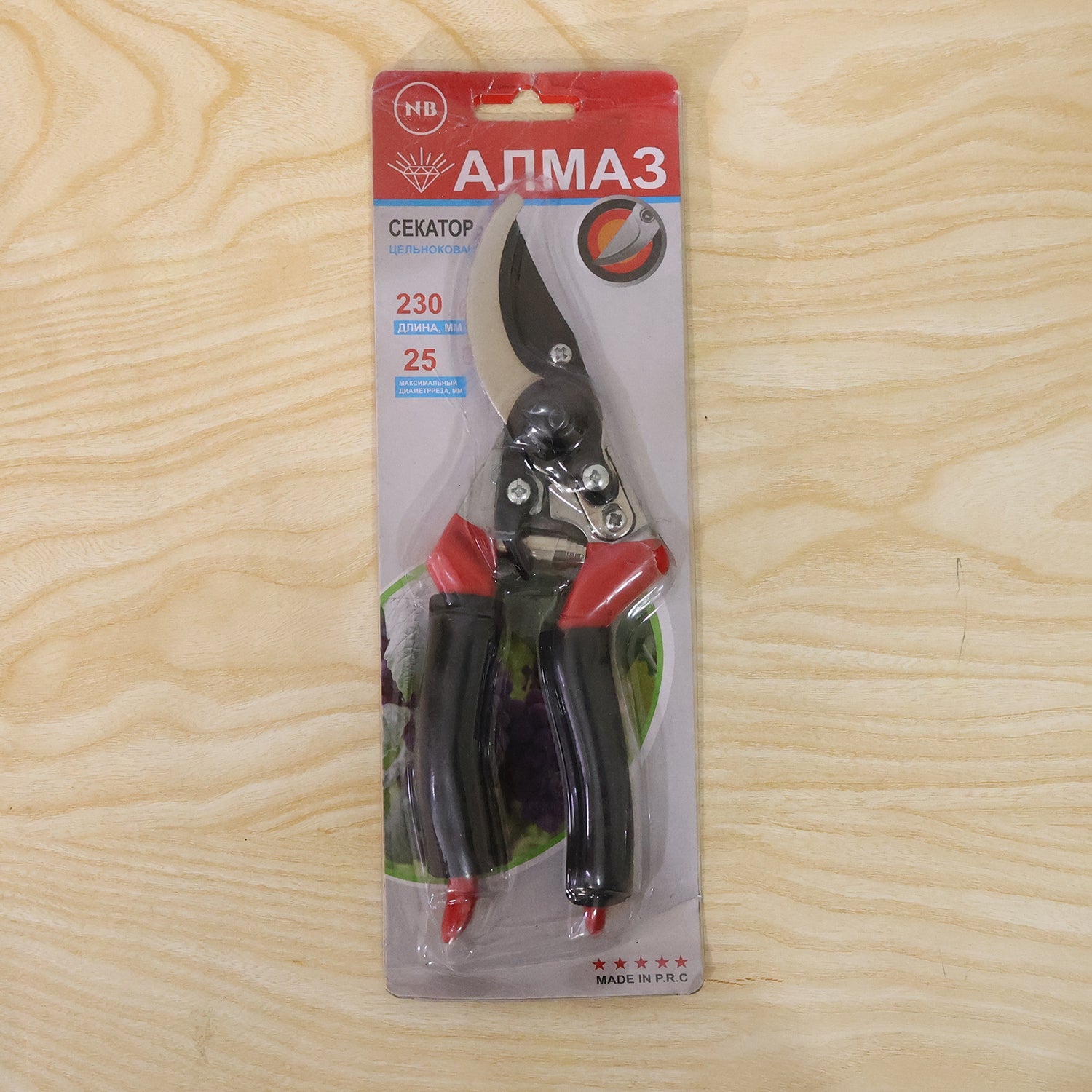 Red and black garden pruners in clear plastic and red cardboard packaging on a light wood background.