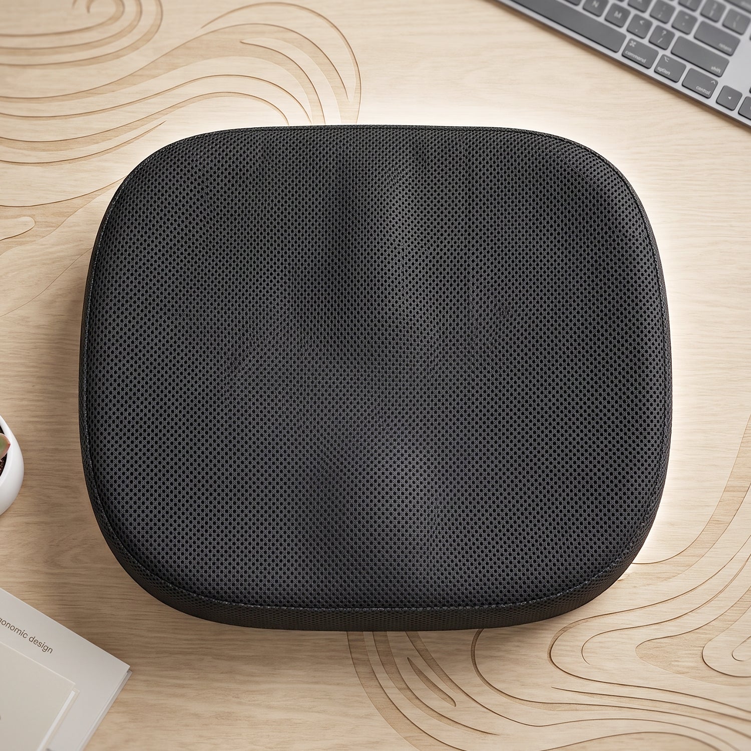 Top-down view of a black perforated ergonomic seat cushion on a light wood desk with a keyboard.