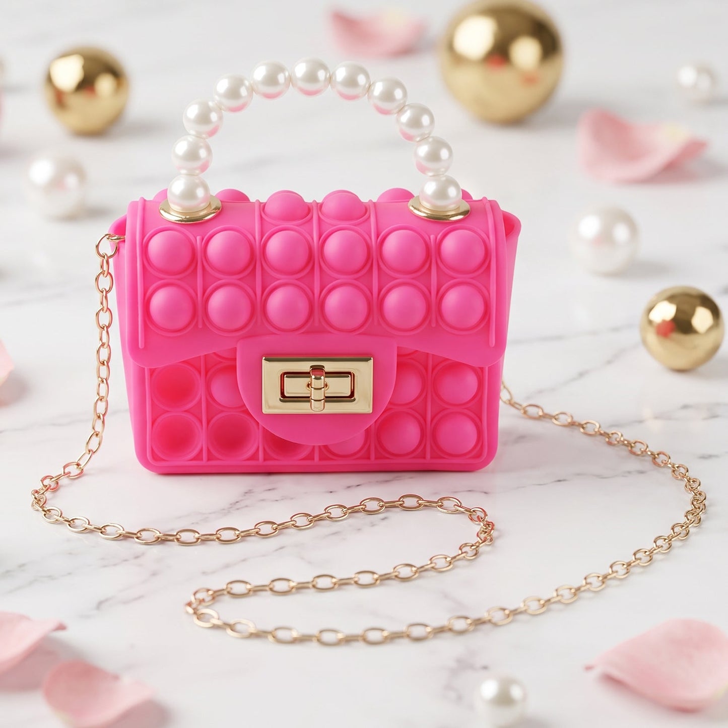 A pink pop-it purse with a pearl handle and gold chain sits on a marble surface, surrounded by gold spheres and pink petals.