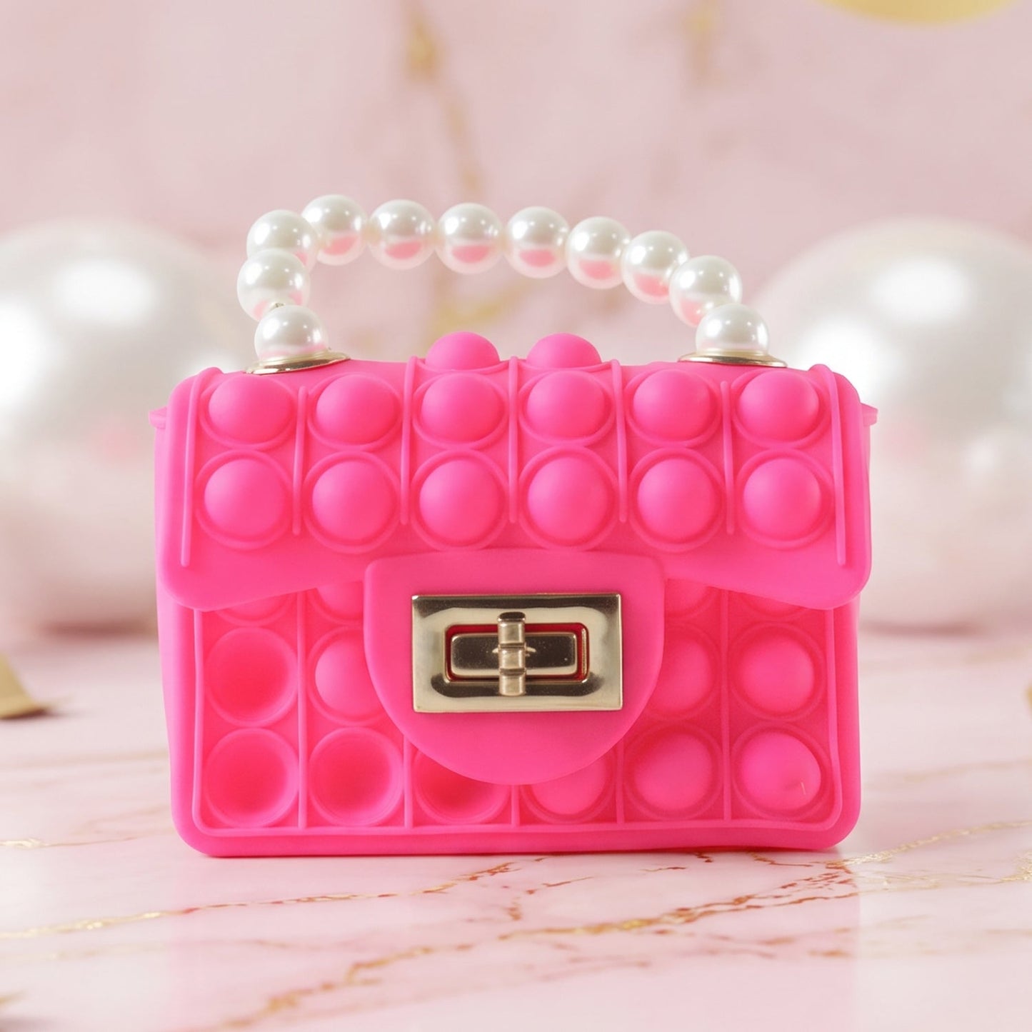 A vibrant pink pop-it purse with a pearl handle and gold clasp sits on a marble surface with blurred white and gold