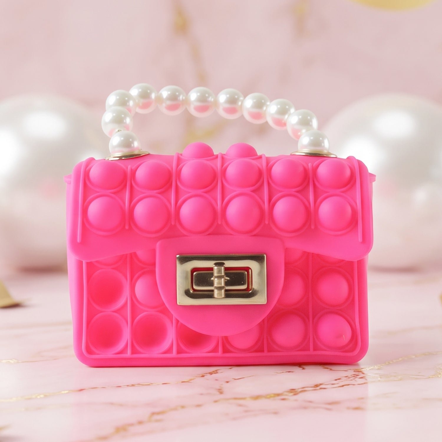 A vibrant pink pop-it purse with a pearl handle and gold clasp sits on a marble surface with blurred white and gold