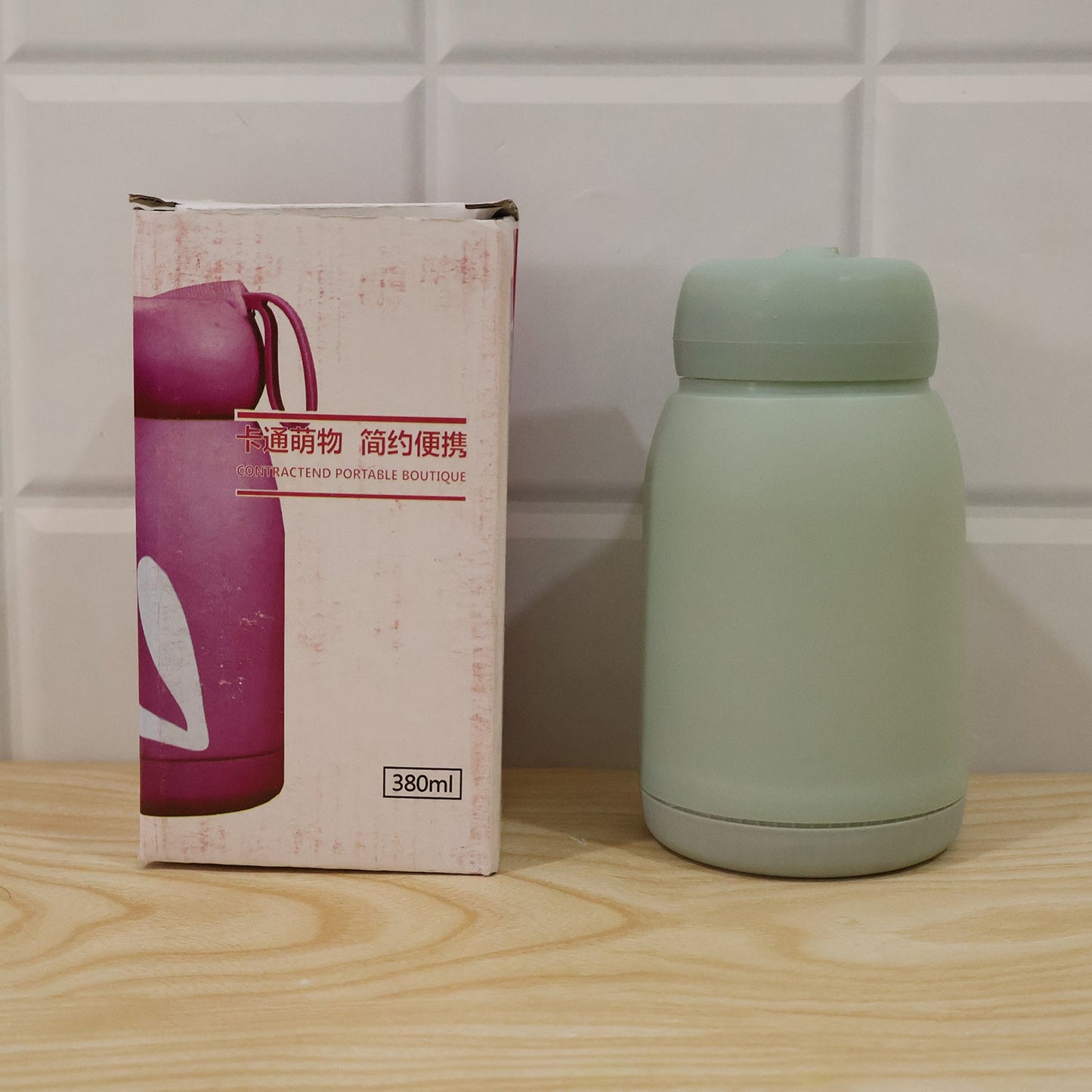 A mint green glass bottle with a bunny print stands next to its pink and white product box on a wooden surface.