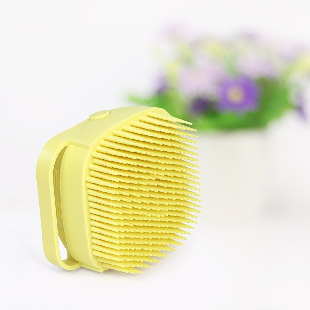 A yellow silicone bath brush is angled to show its bristles, with a blurred white pot of purple flowers in the background.