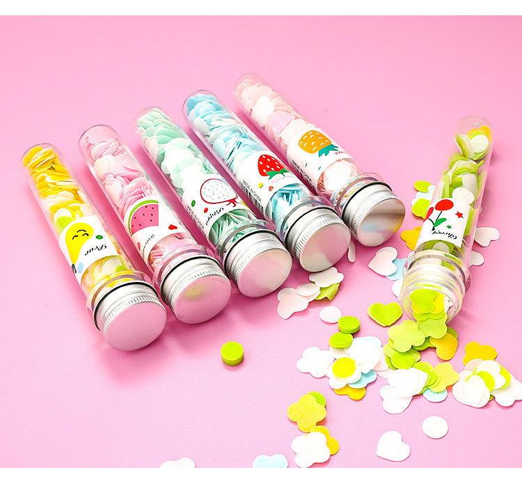 Five clear tubes of colorful flower-shaped paper soap strips with fruit labels are arranged on a pink surface with loose