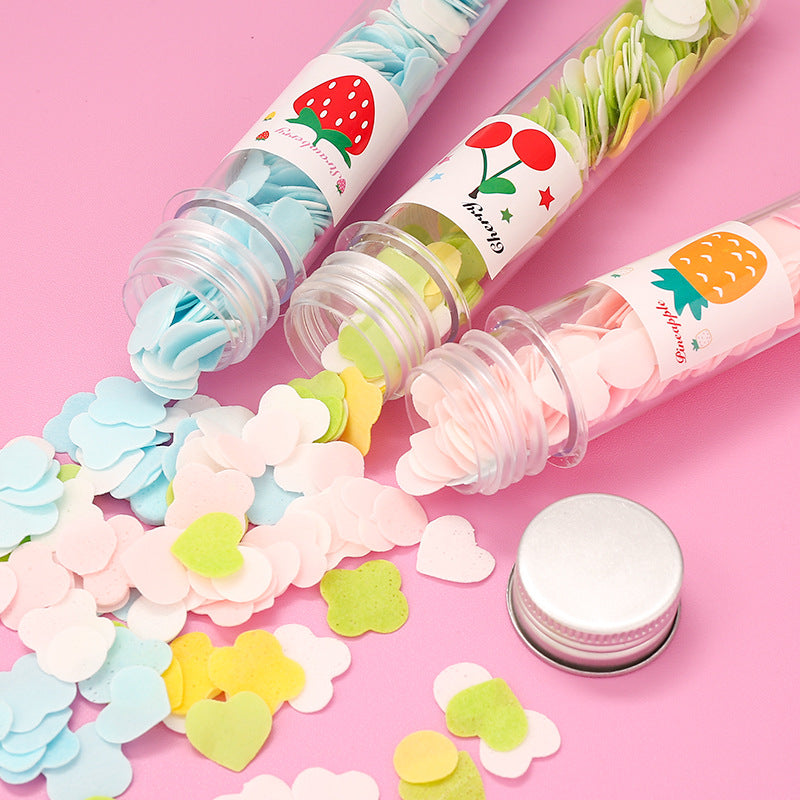 Three clear tubes of colorful flower-shaped paper soap strips are spilling out onto a pink surface, with a silver cap nearby.