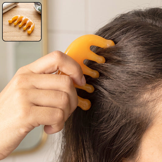 A person's hand holds an orange scalp massager comb to their dark hair; a smaller inset shows the comb on a wooden surface.
