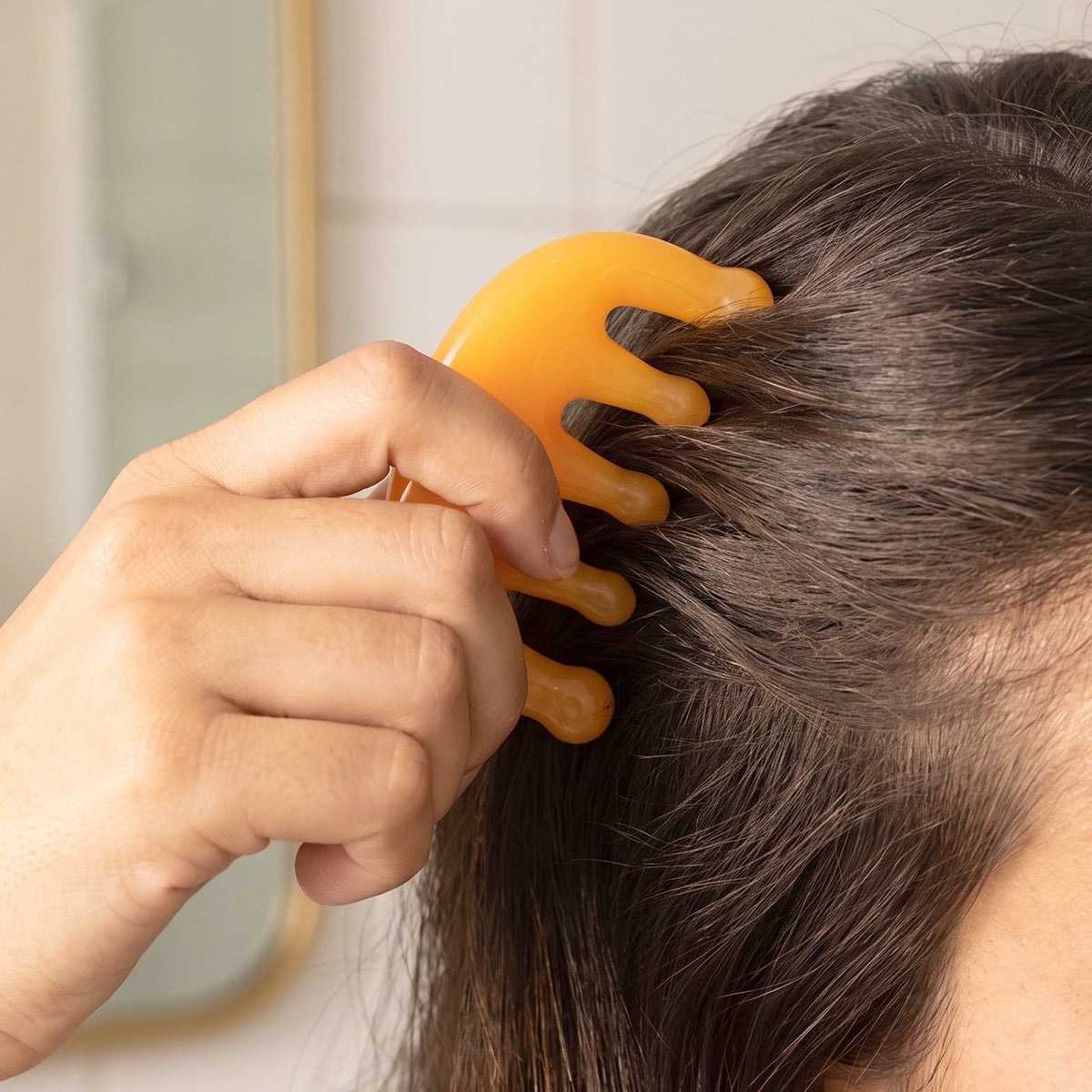 A hand holds the translucent orange scalp massager comb against dark hair, with a blurred mirror reflection in the