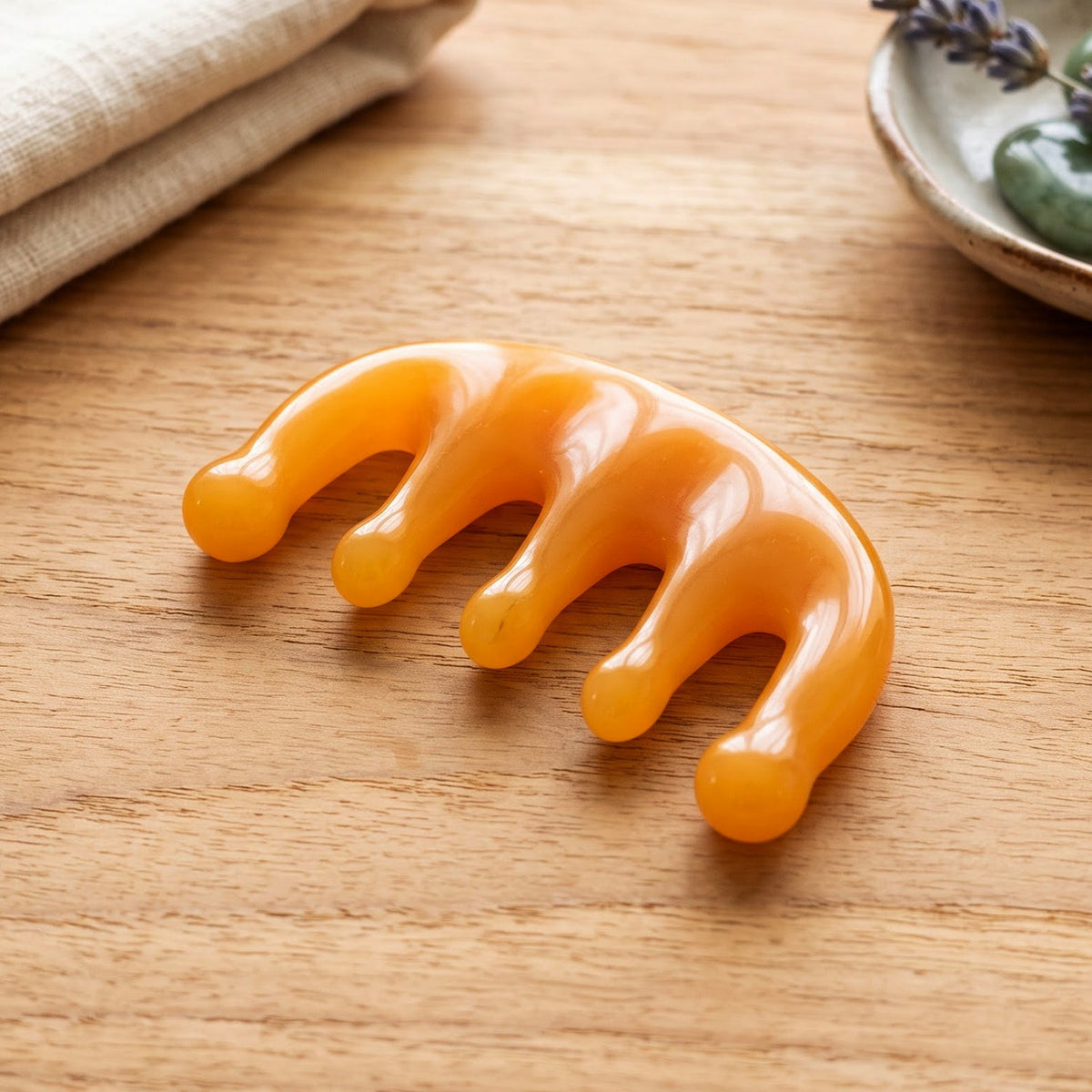 The orange scalp massager comb rests on a light wooden surface next to a folded towel and a small dish with lavender.