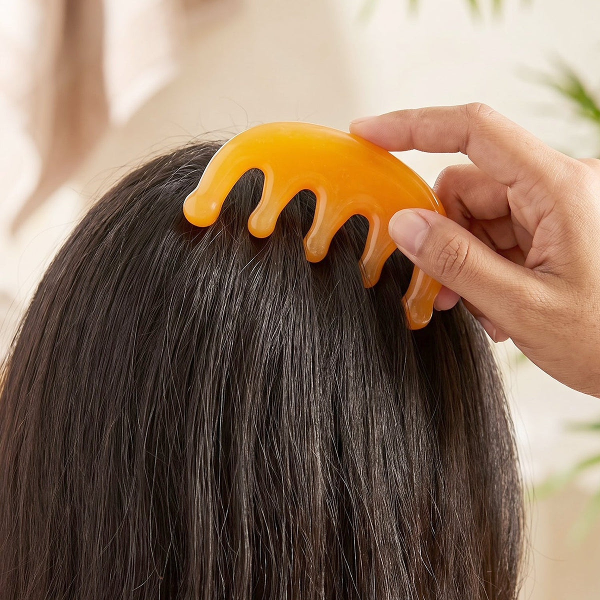 A hand gently presses the orange scalp massager comb onto the back of a person's dark head.